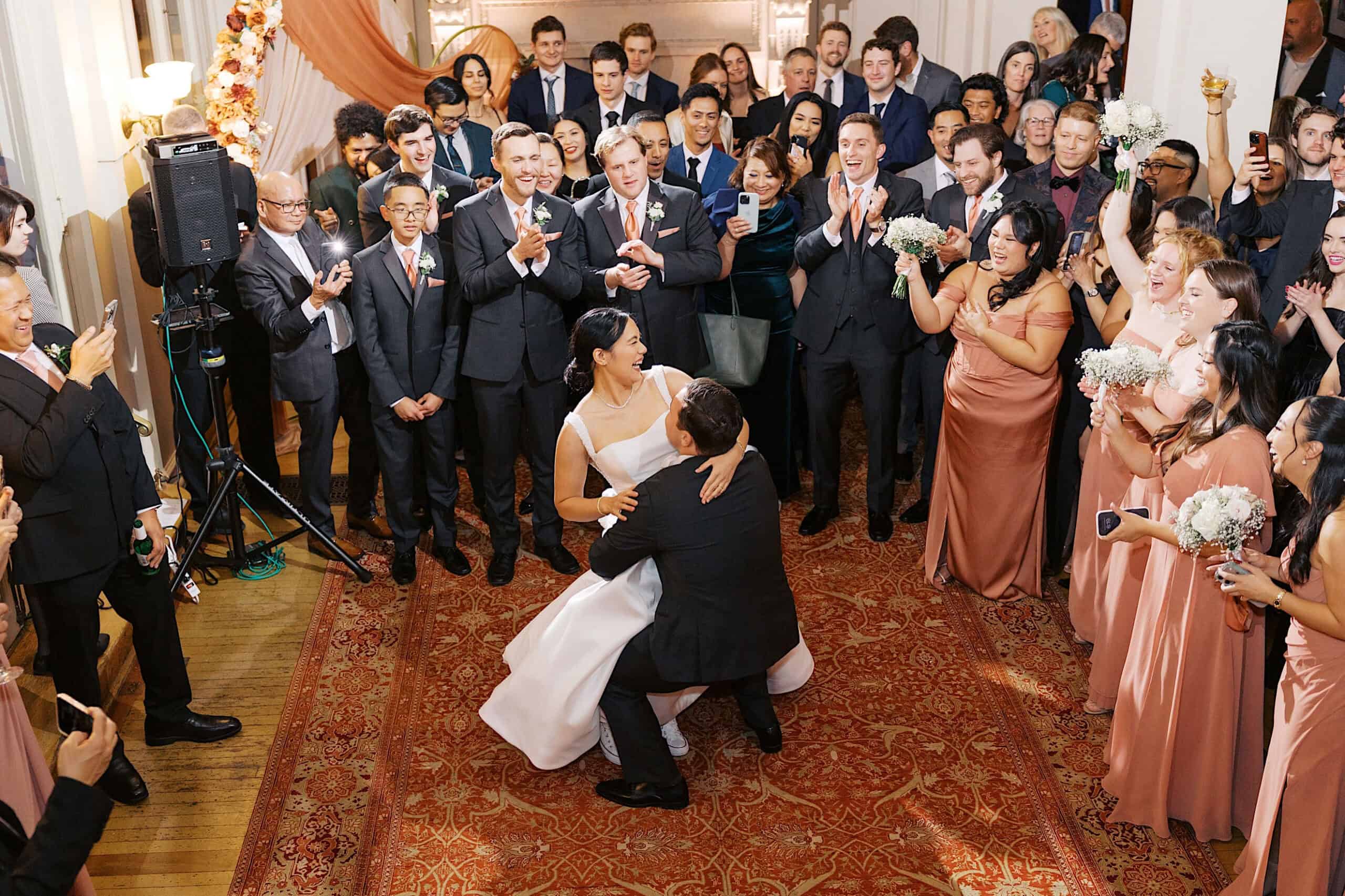 A bride and groom share a dance in the center of a crowd, with bridesmaids in peach dresses and guests watching and taking photos at an elegant Cairnwood Estate Wedding.