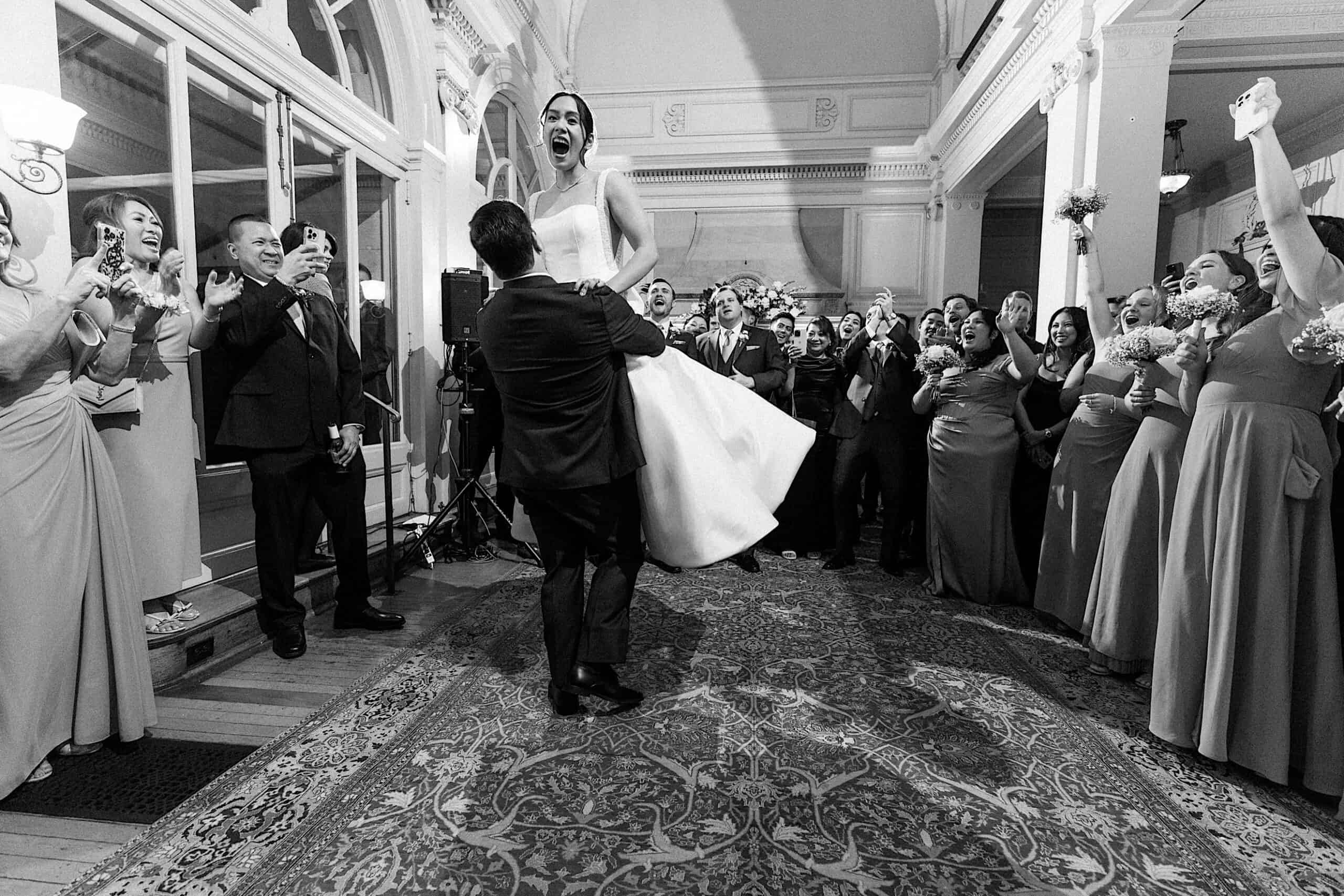A groom lifts a bride in a white dress as guests cheer and clap around them during an elegant Cairnwood Estate Wedding reception.
