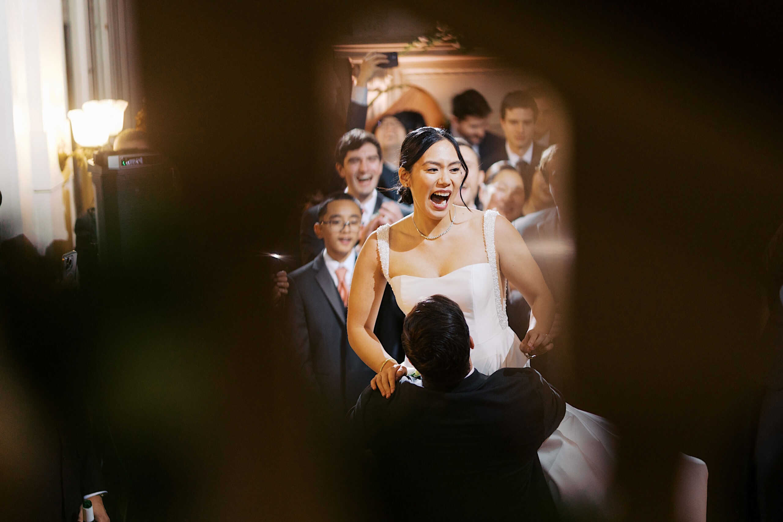 A bride in a white dress is lifted on a chair during an elegant Cairnwood Estate Wedding, surrounded by smiling and cheering guests.