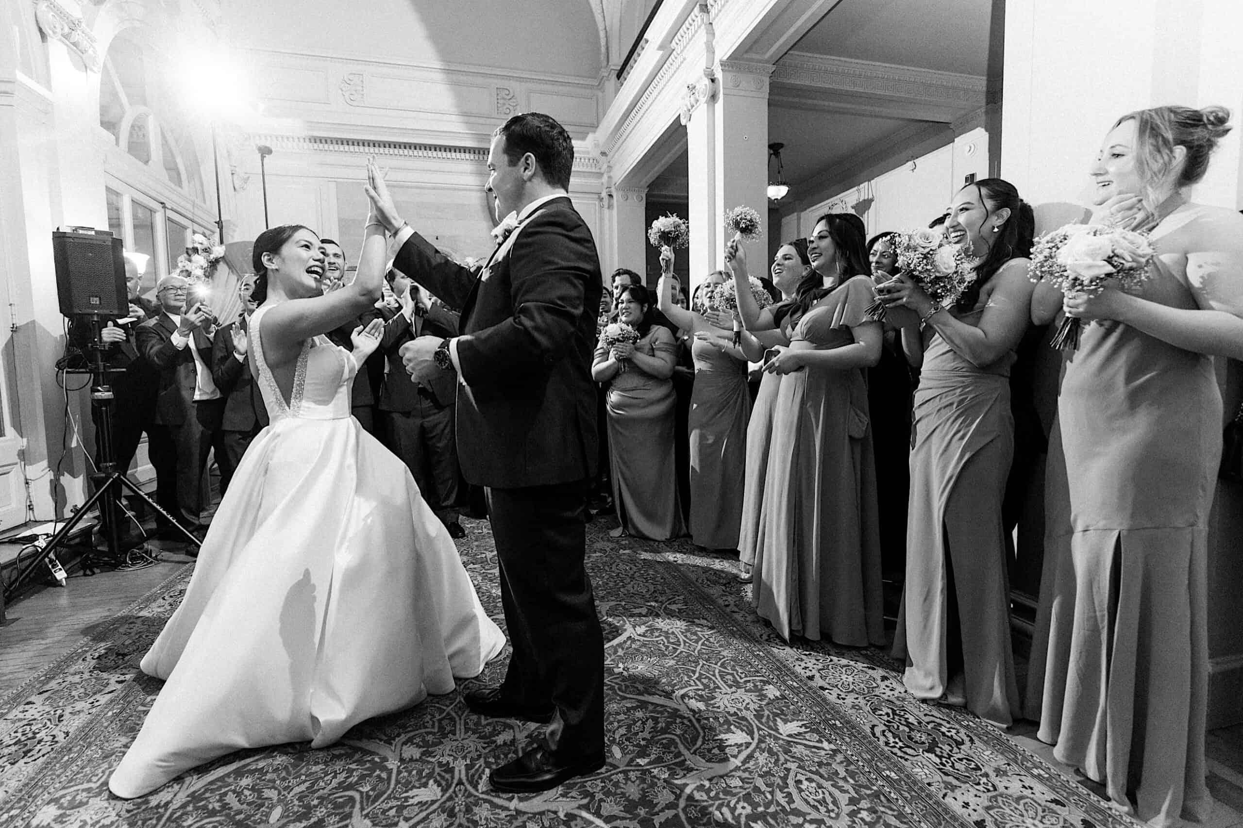 A bride and groom high-five on the dance floor at their elegant Cairnwood Estate Wedding, surrounded by smiling bridesmaids holding bouquets in a beautifully decorated indoor venue.
