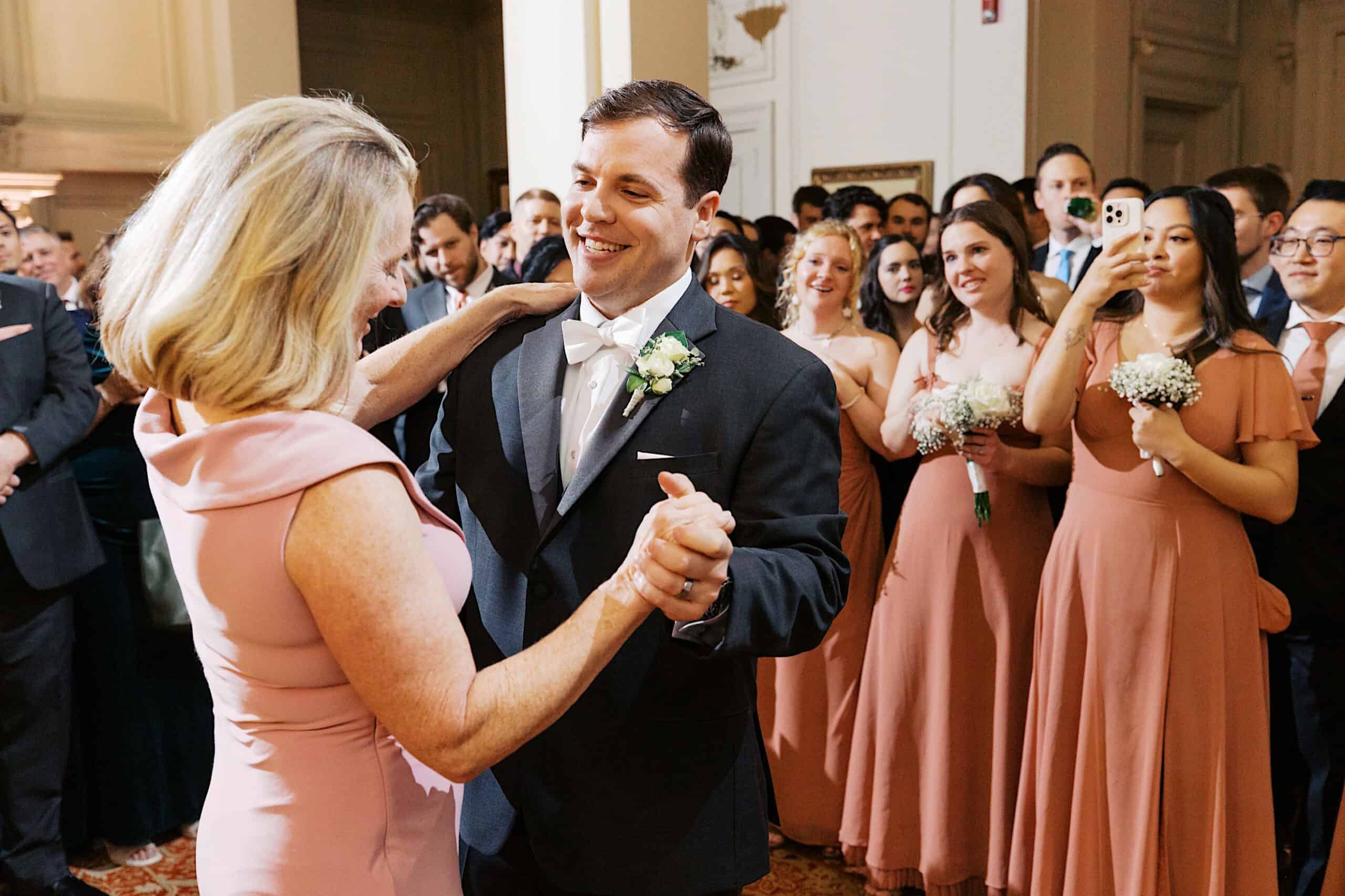 A man in a tuxedo dances with a woman in a pink dress as guests and bridesmaids watch and take photos at this elegant Cairnwood Estate Wedding.