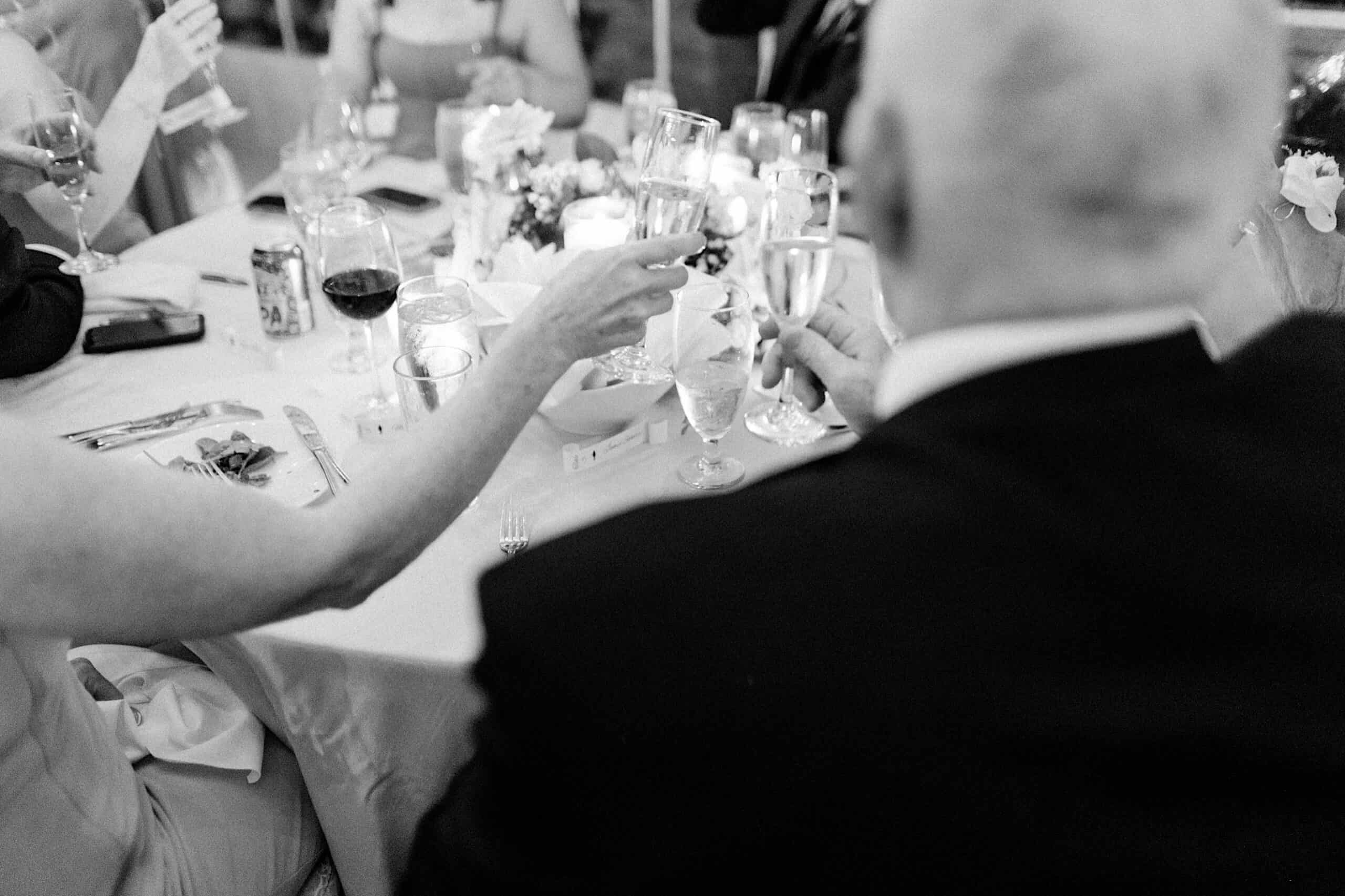 People seated at a table raise glasses in a toast; the table is set with drinks, utensils, and a centerpiece. This black-and-white photo captures the joy of an elegant Cairnwood Estate Wedding celebration.