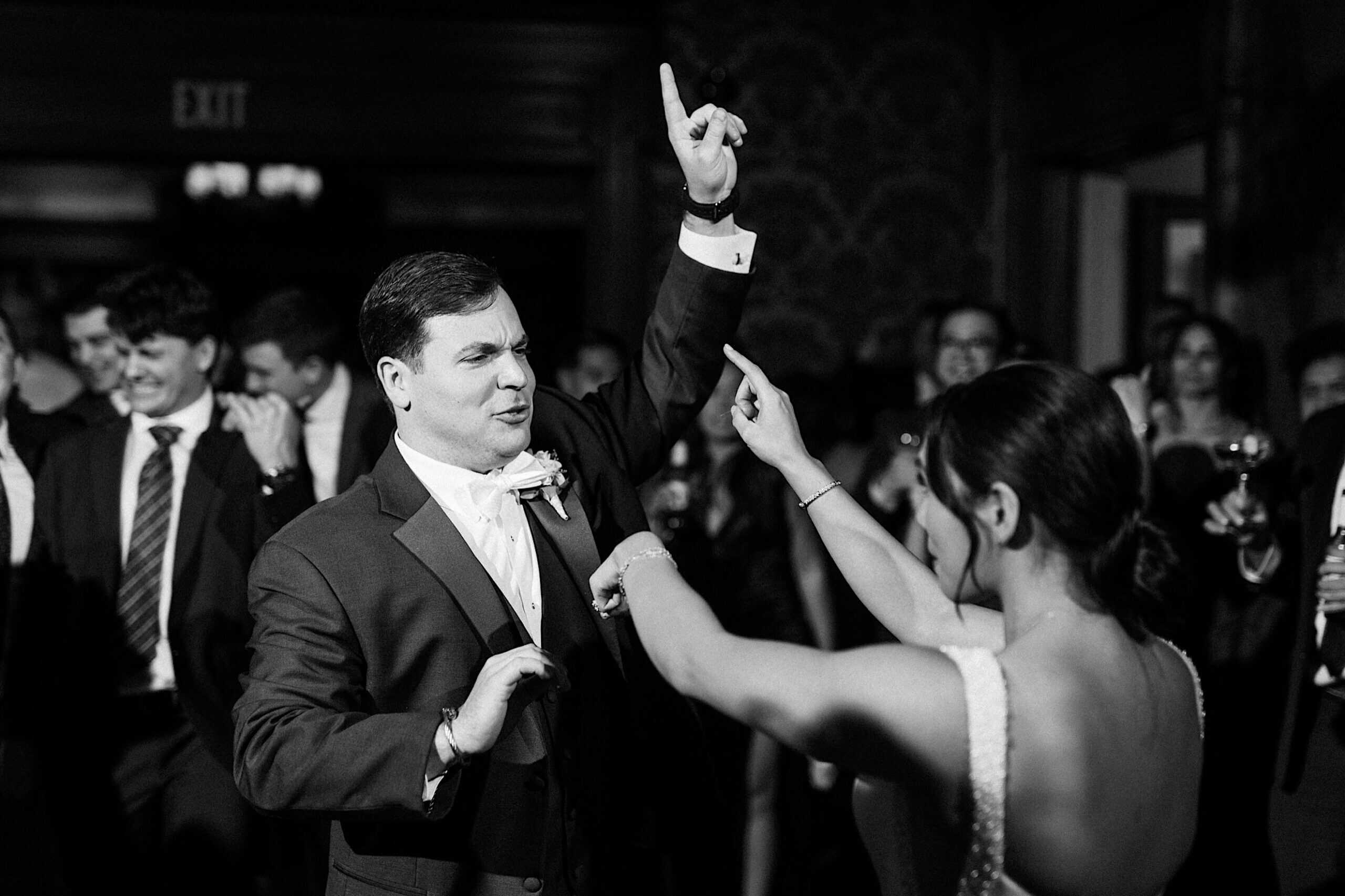 A man in a suit and a woman in a dress dance together at an elegant Cairnwood Estate Wedding, both raising one arm and pointing upward, with other guests visible in the background.