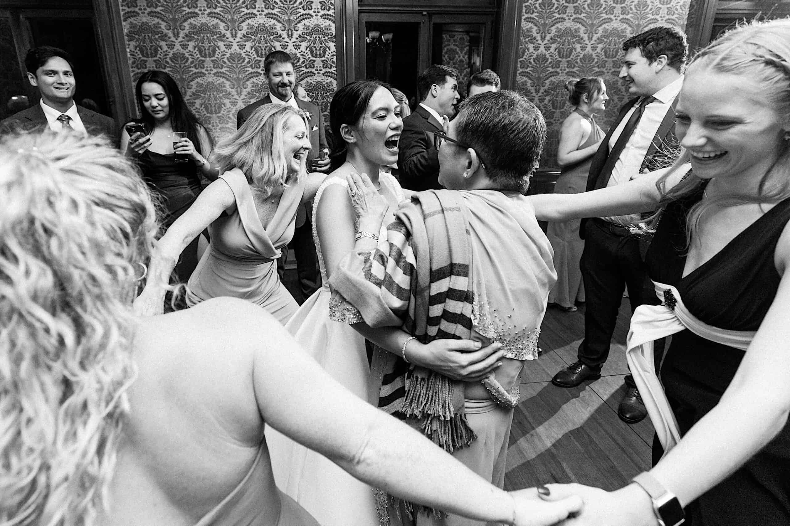 A group of people, dressed formally, hold hands and dance in a circle at an elegant Cairnwood Estate Wedding, smiling and laughing at the indoor event.