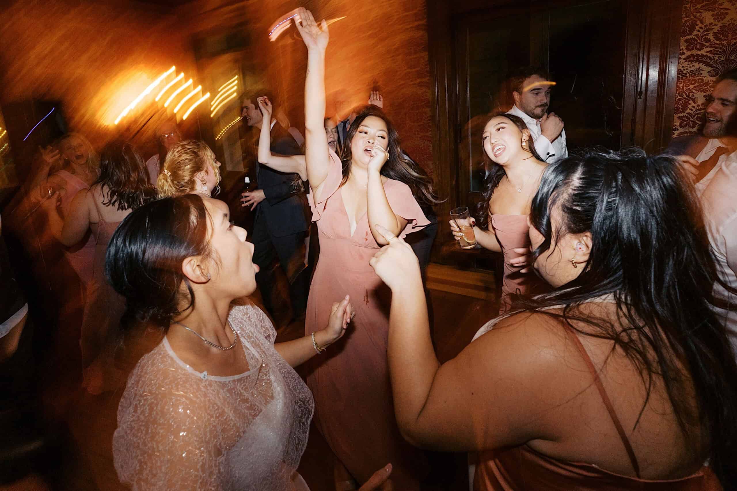 A group of people in formal attire dance energetically at an elegant Cairnwood Estate Wedding, some with drinks in hand, in a warmly lit room with blurred motion.