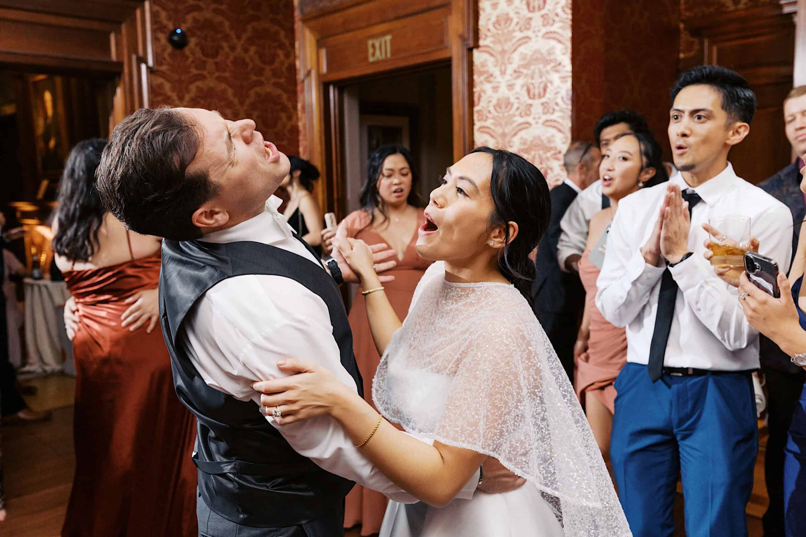 A bride and groom sing together on the dance floor at their elegant Cairnwood Estate Wedding, surrounded by cheering and clapping wedding guests in formal attire.