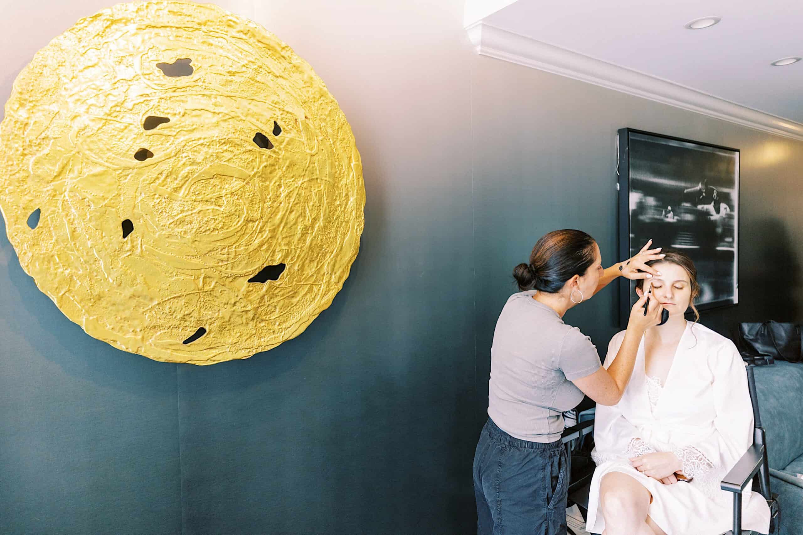 A makeup artist applies makeup to a seated woman in a white robe beside a large yellow circular wall sculpture before her Franklin Institute wedding.