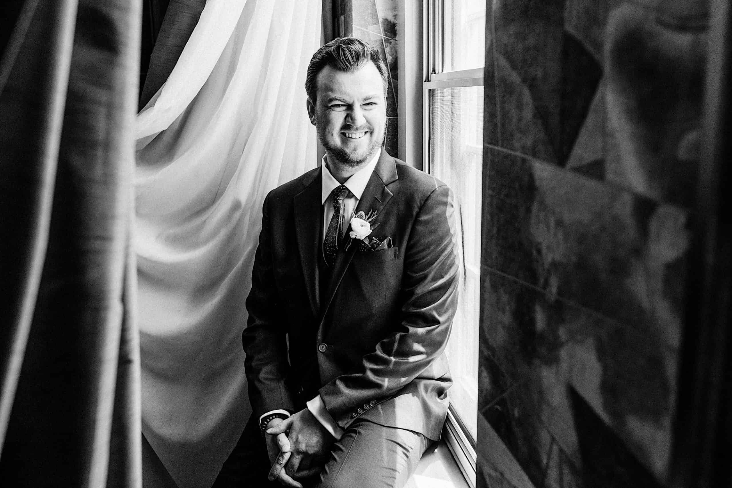 A man in a suit with a boutonnière sits on a windowsill, smiling, with curtains and natural light in the background at a Franklin Institute wedding. Black and white photo.