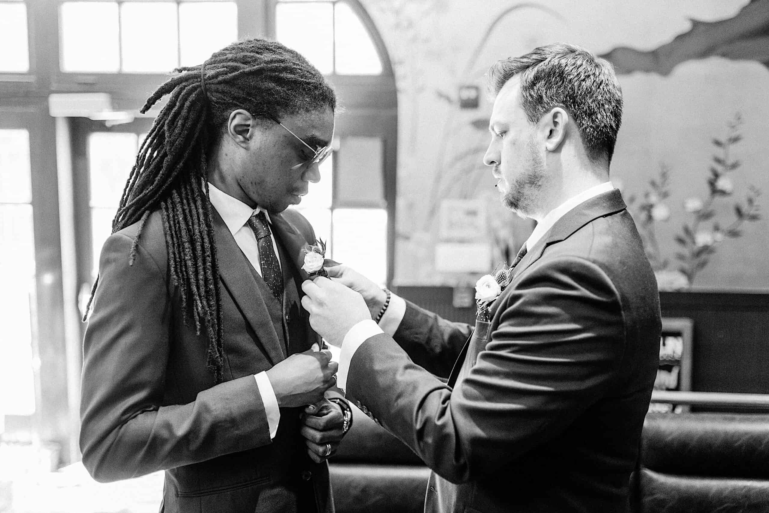 Two men in suits stand indoors; one adjusts the other's boutonniere. They appear focused and are dressed formally, possibly preparing for a Franklin Institute wedding or another special event.