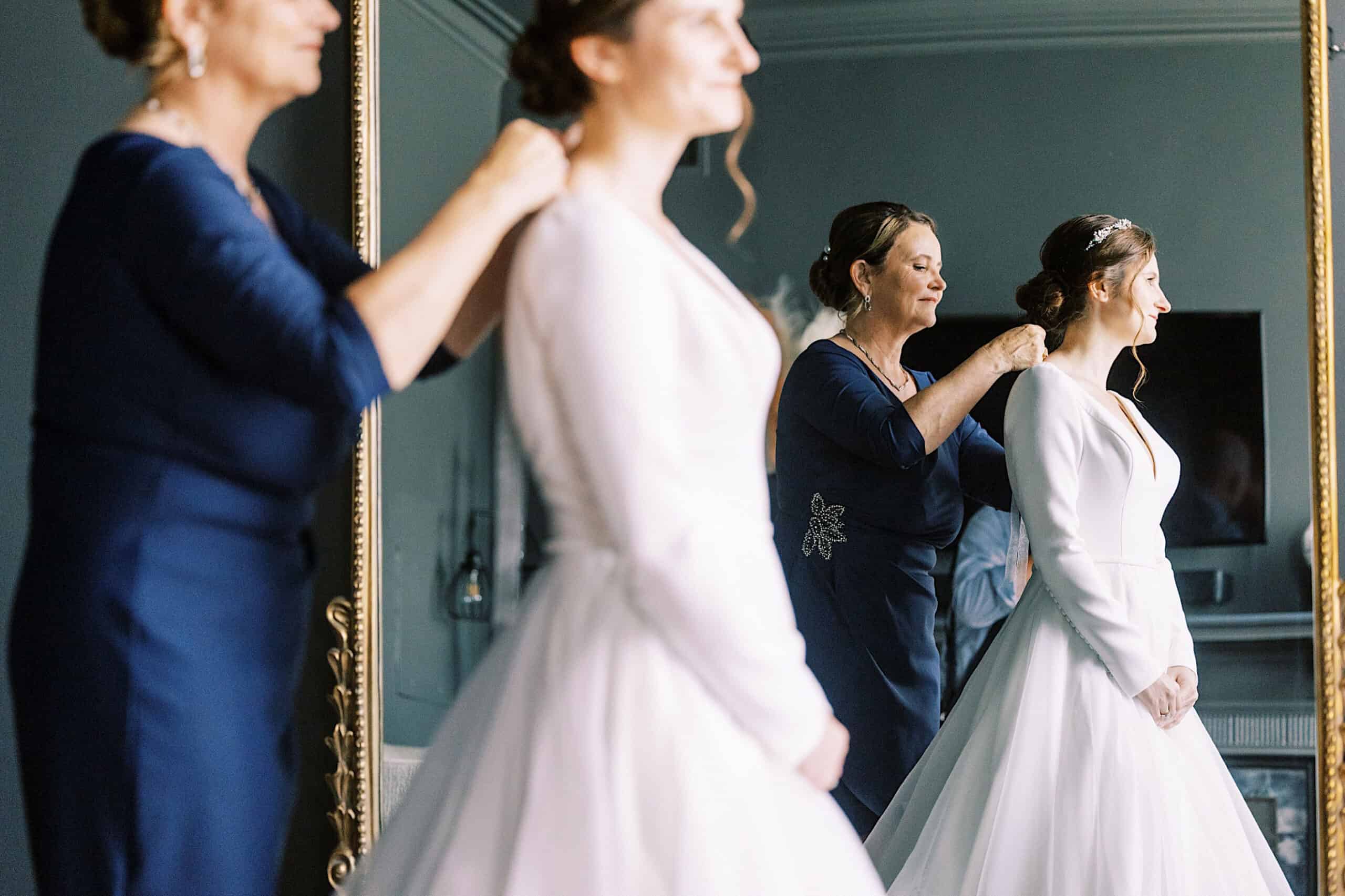 Two women in blue dresses help a bride adjust her necklace in front of a large mirror at her Franklin Institute wedding. The bride, wearing a white gown, stands facing the mirror as final touches are made before the celebration.