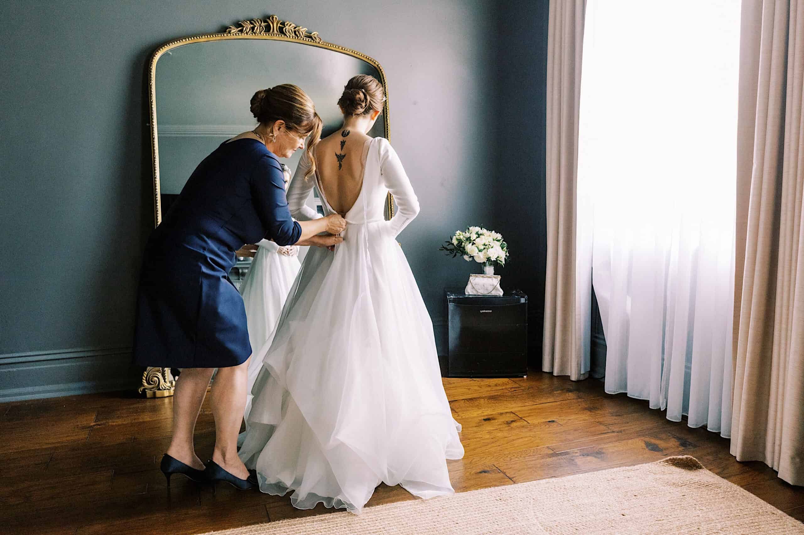 A woman in a navy dress helps a bride in a white gown button up her dress in a room with a large mirror and white curtains before their elegant Franklin Institute wedding.