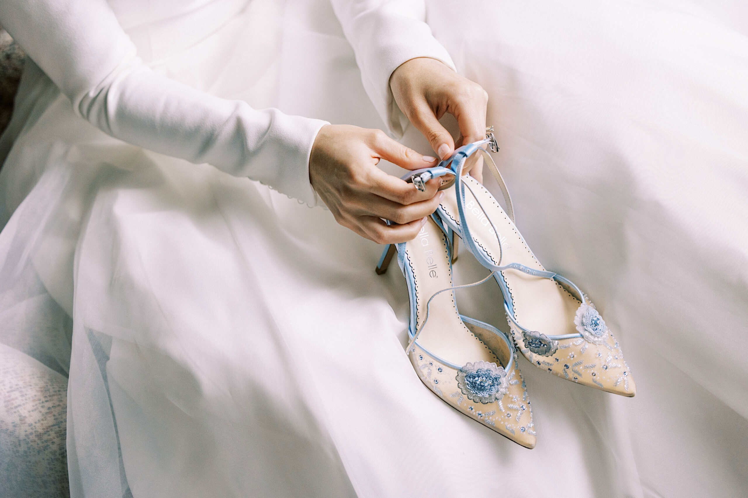 A person in a white dress holds a pair of pointed-toe, floral-embroidered high heels with blue trim and decorative flowers, perfect for a romantic Franklin Institute wedding.