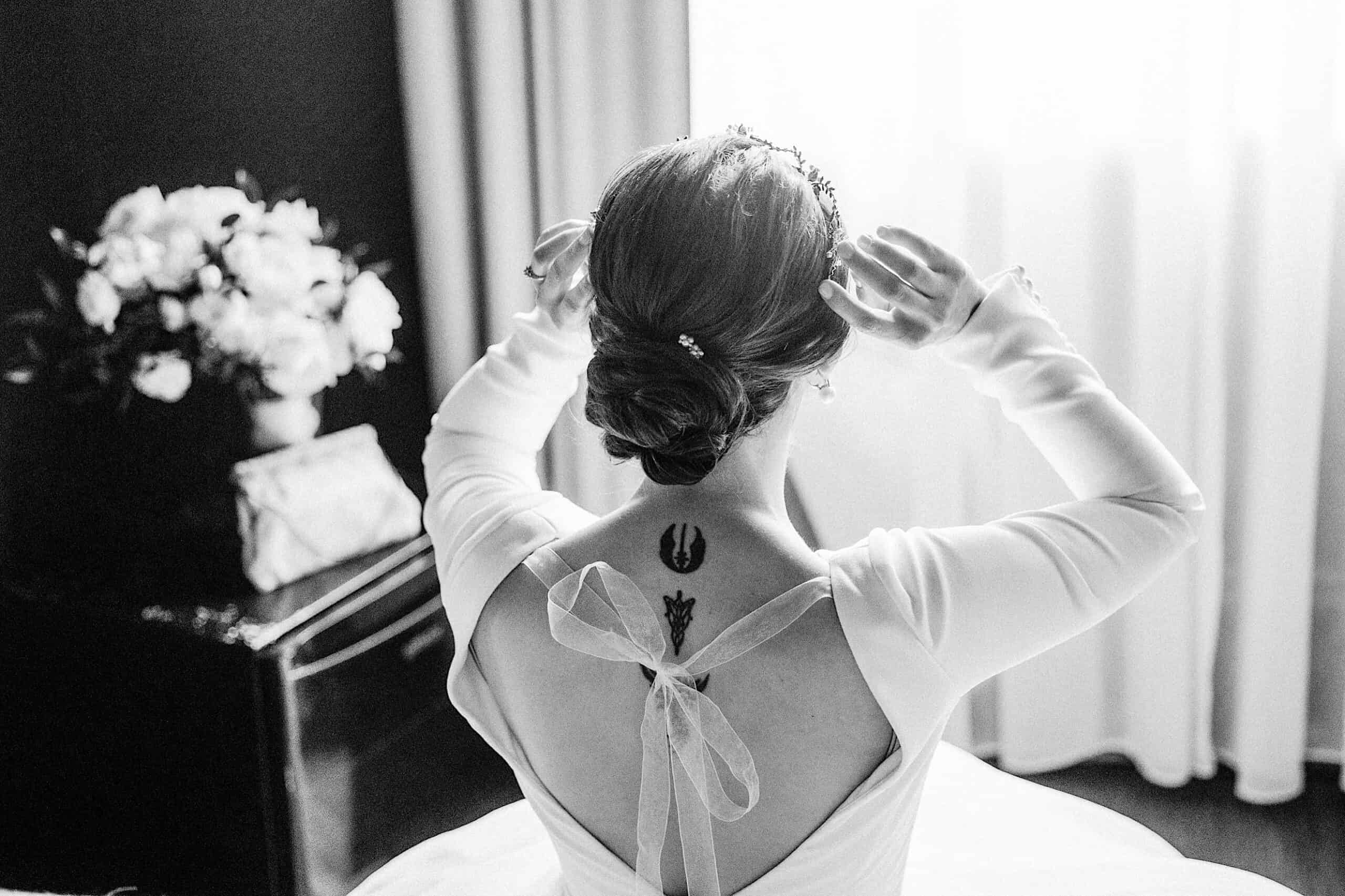 A woman in a white dress with a bow on the back adjusts her hair, revealing two tattoos on her upper back, while sitting near a table with a bouquet of flowers at her elegant Franklin Institute wedding.