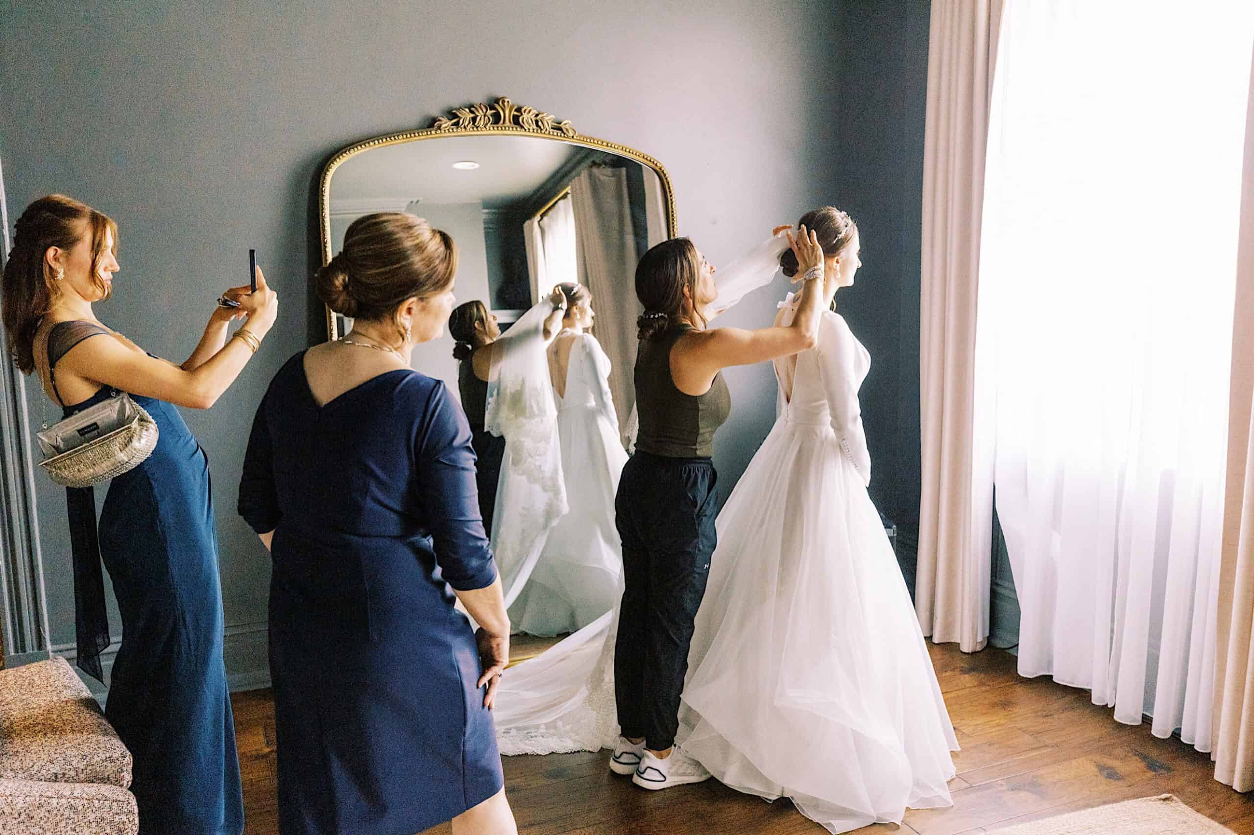 A bride stands by a window in her white dress as a stylist perfects her hair; two women watch, one snapping a photo, with a large mirror reflecting the scene at this elegant Franklin Institute wedding.