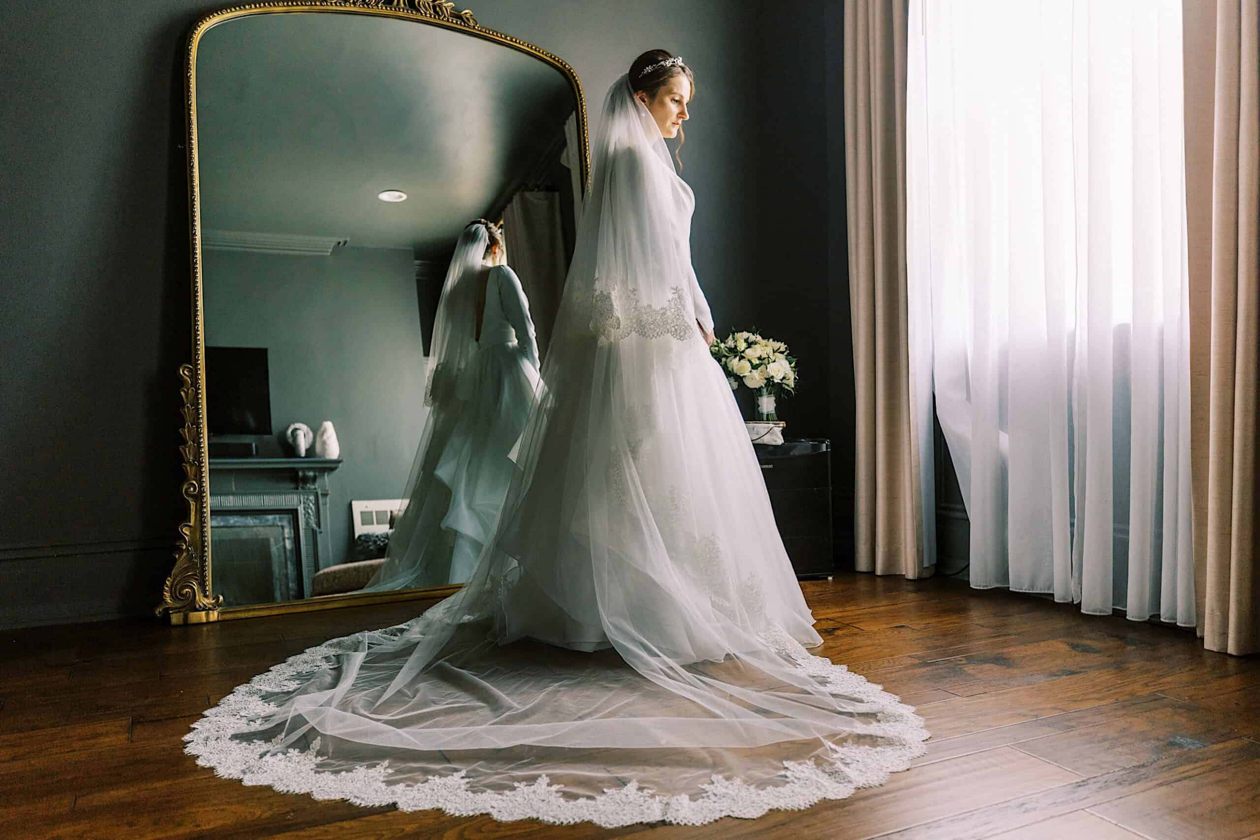 A bride in a white wedding gown and veil stands near a large mirror in a softly lit room at her Franklin Institute wedding, her reflection visible as sunlight streams through sheer curtains.