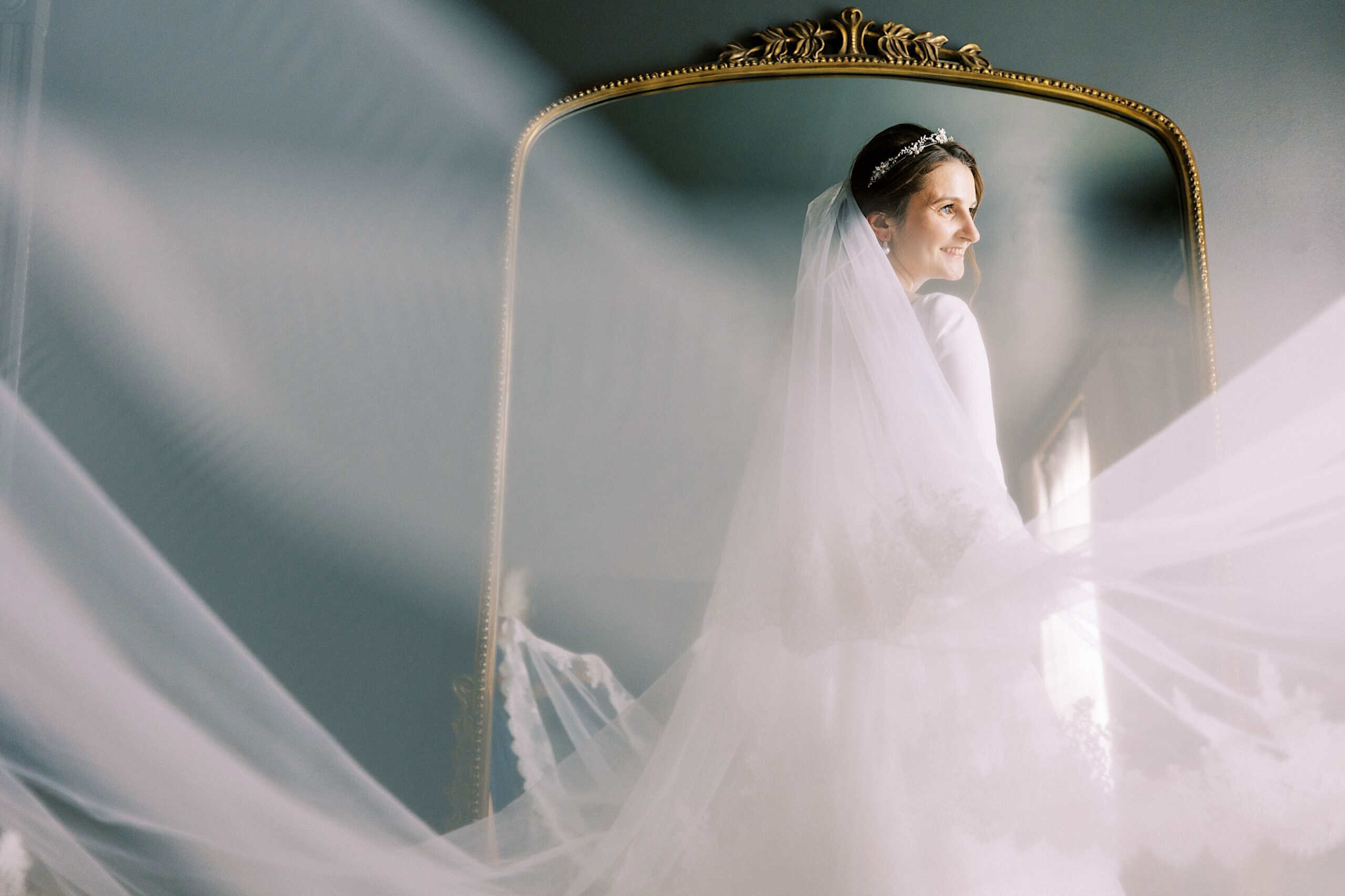 A bride in a white dress and veil stands before a large ornate mirror at her Franklin Institute wedding, her veil flowing around her as soft light streams in from the side.