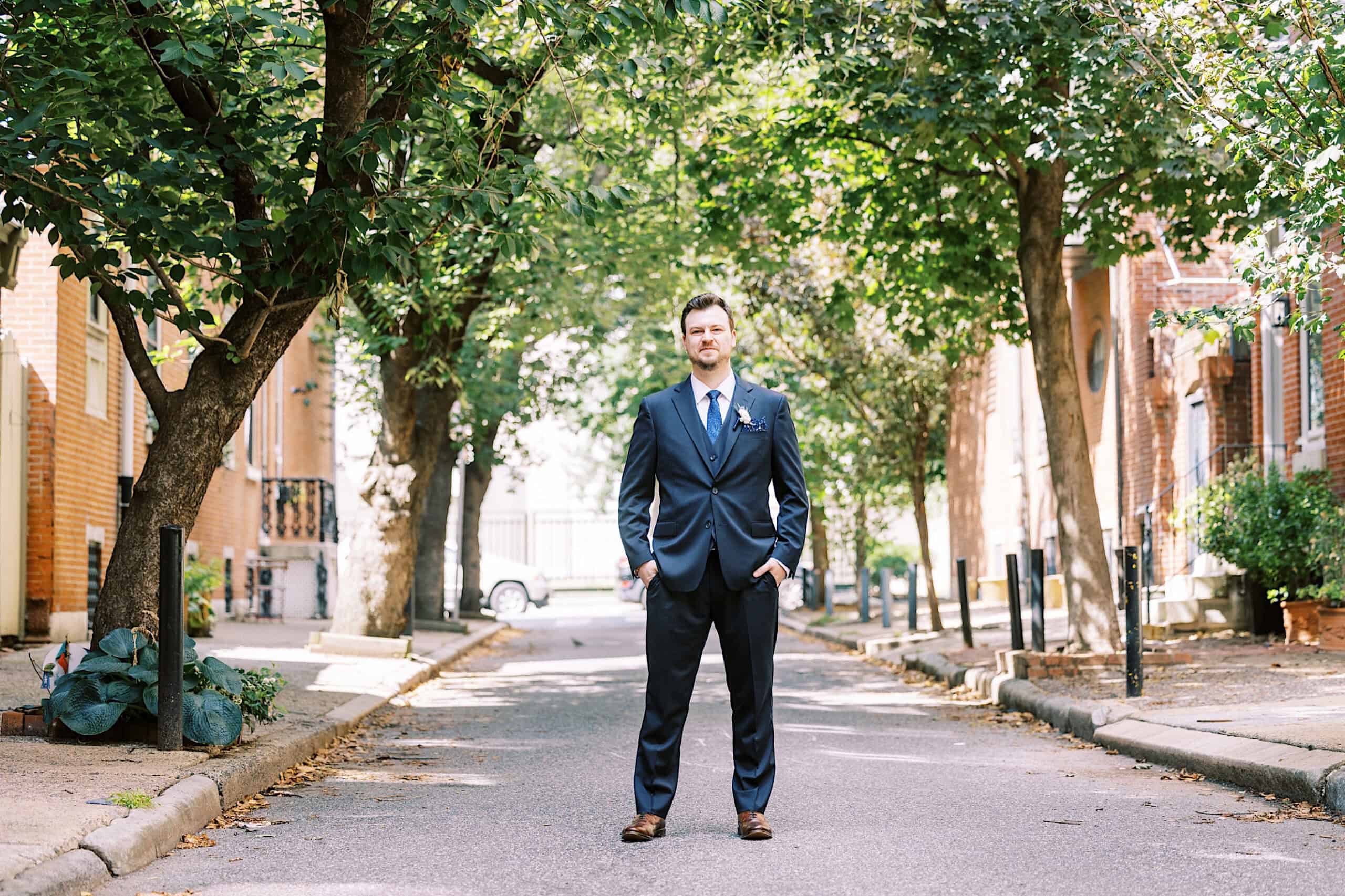 A man in a dark suit stands in the middle of a tree-lined residential street, facing the camera with hands in his pockets, as if on his way to a Franklin Institute wedding.