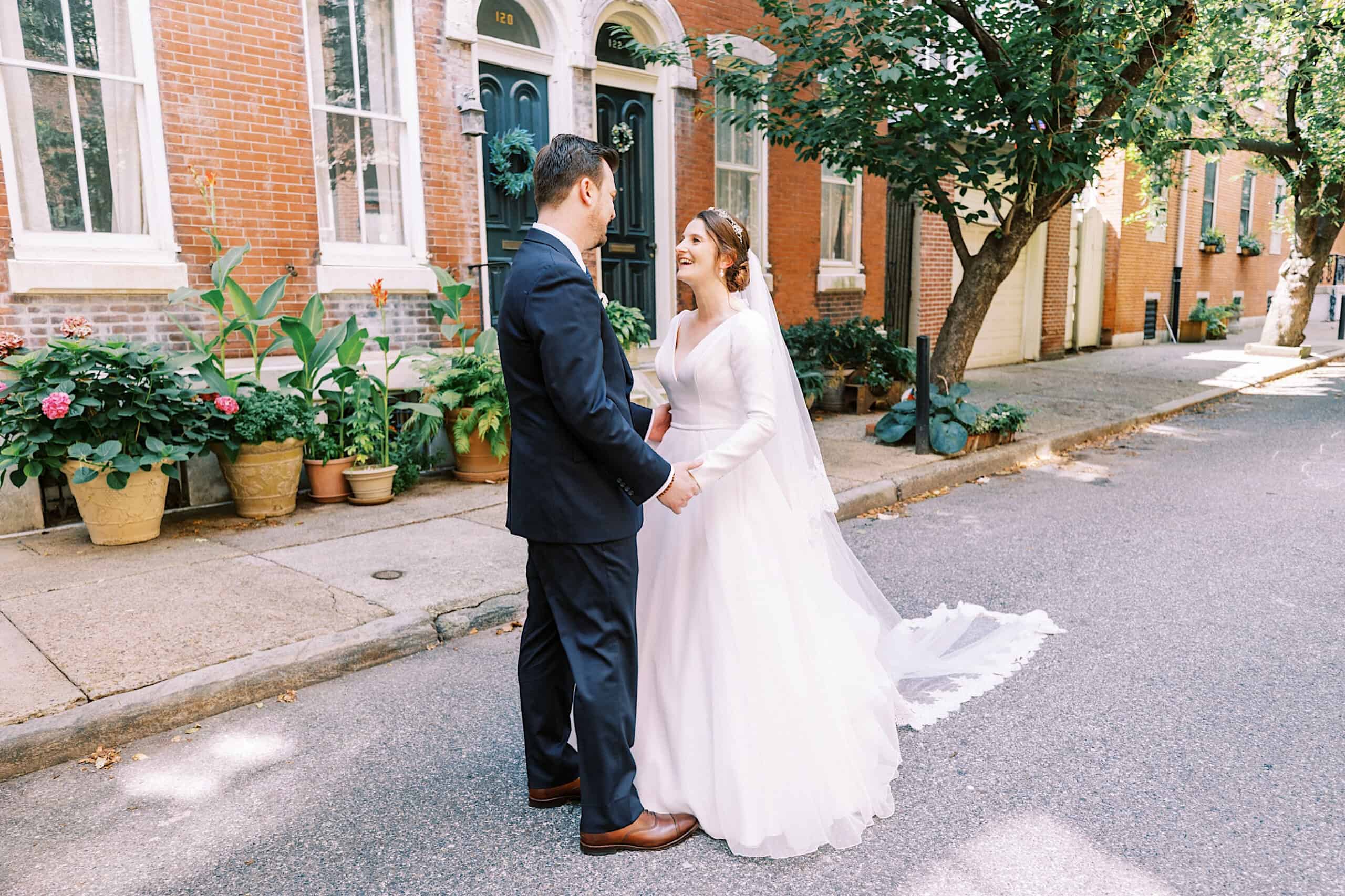 A bride and groom stand facing each other on a quiet, tree-lined street in front of a brick building, dressed in wedding attire after their intimate Franklin Institute wedding.