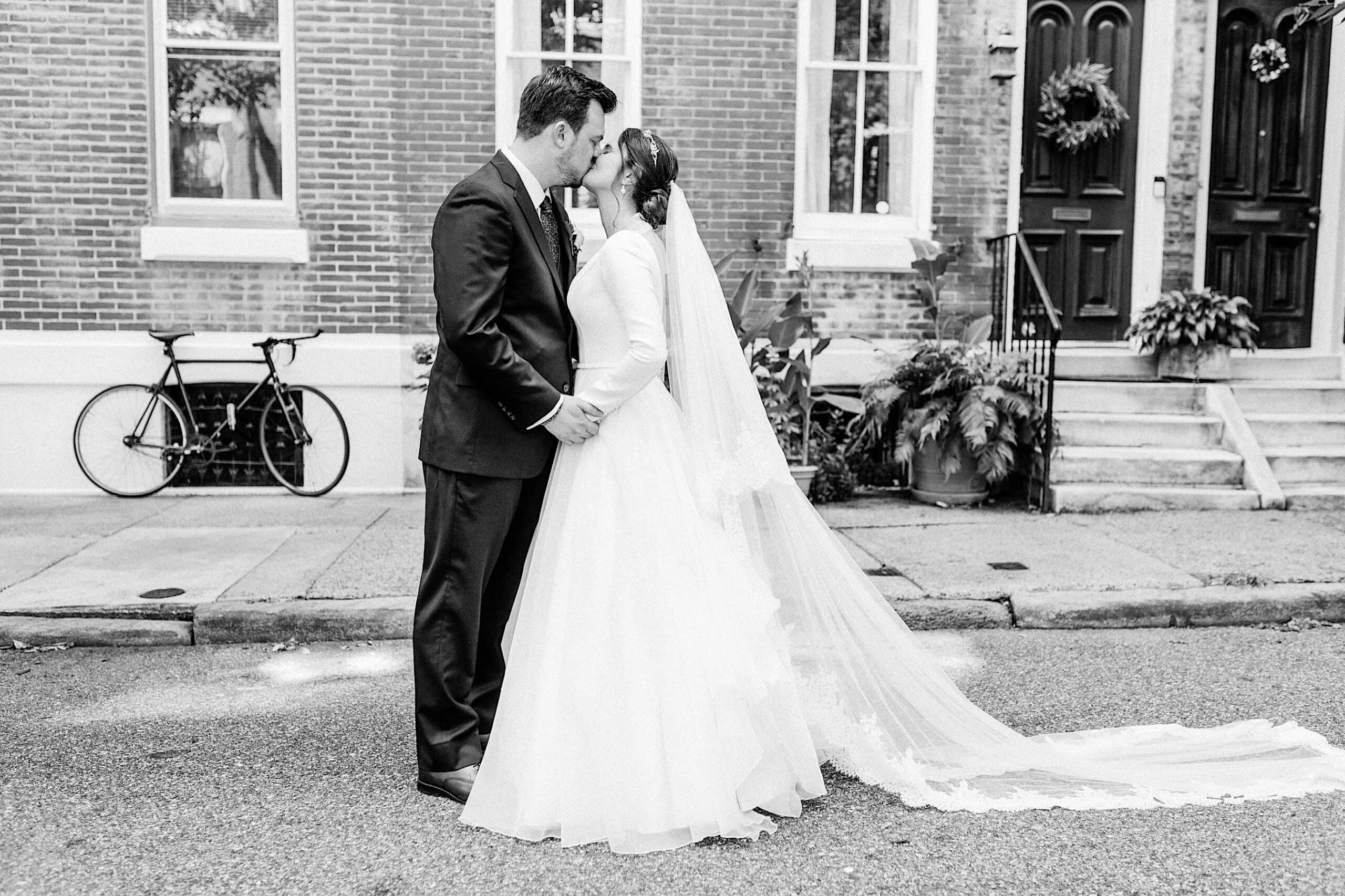 A bride and groom stand in the street, holding hands and kissing, with a brick building, a bicycle, and potted plants in the background after their Franklin Institute wedding.