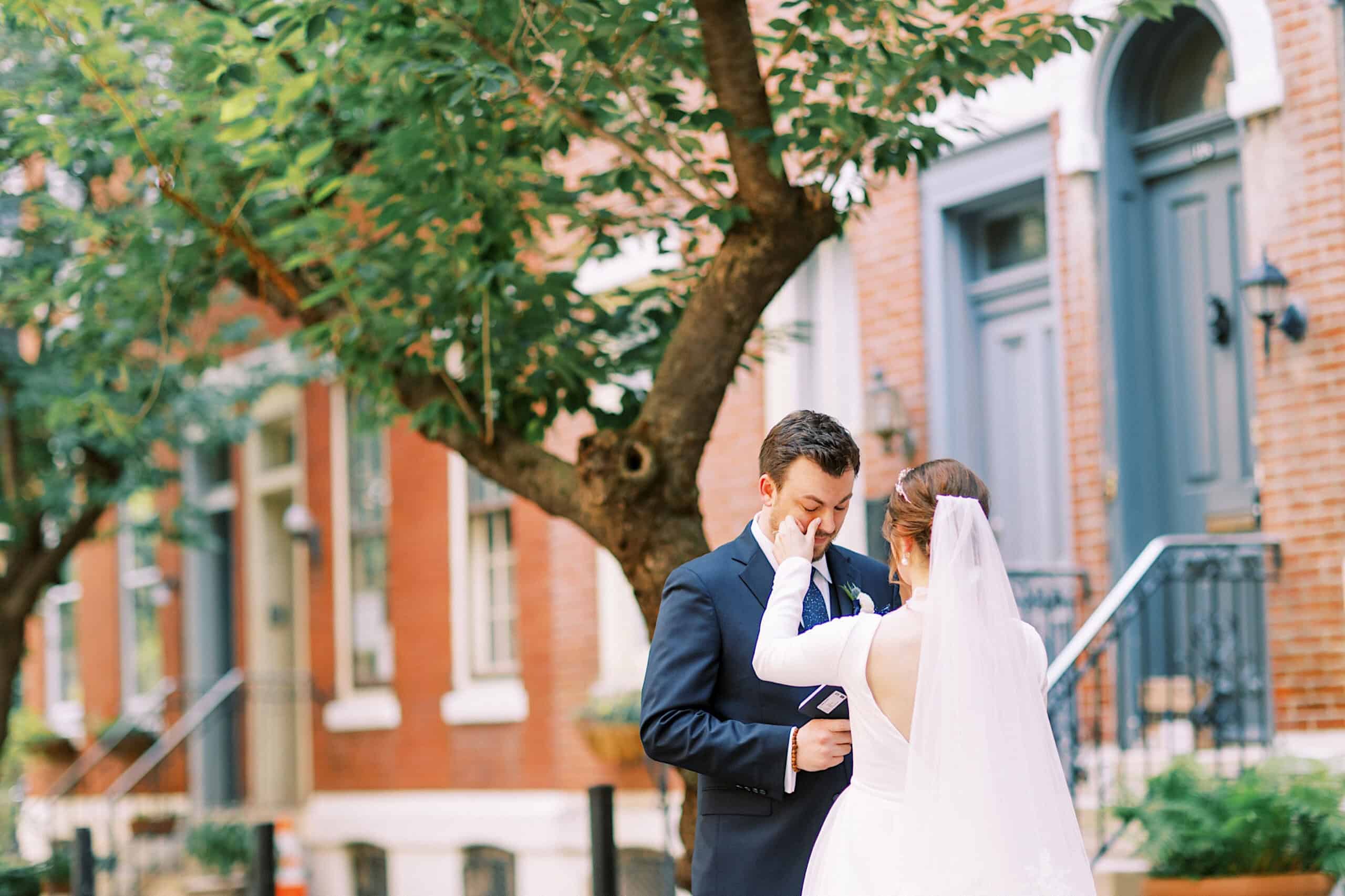 A bride in a white dress gently wipes a tear from the groom’s face as they stand together on a tree-lined city sidewalk, just steps away from their unforgettable Franklin Institute wedding.