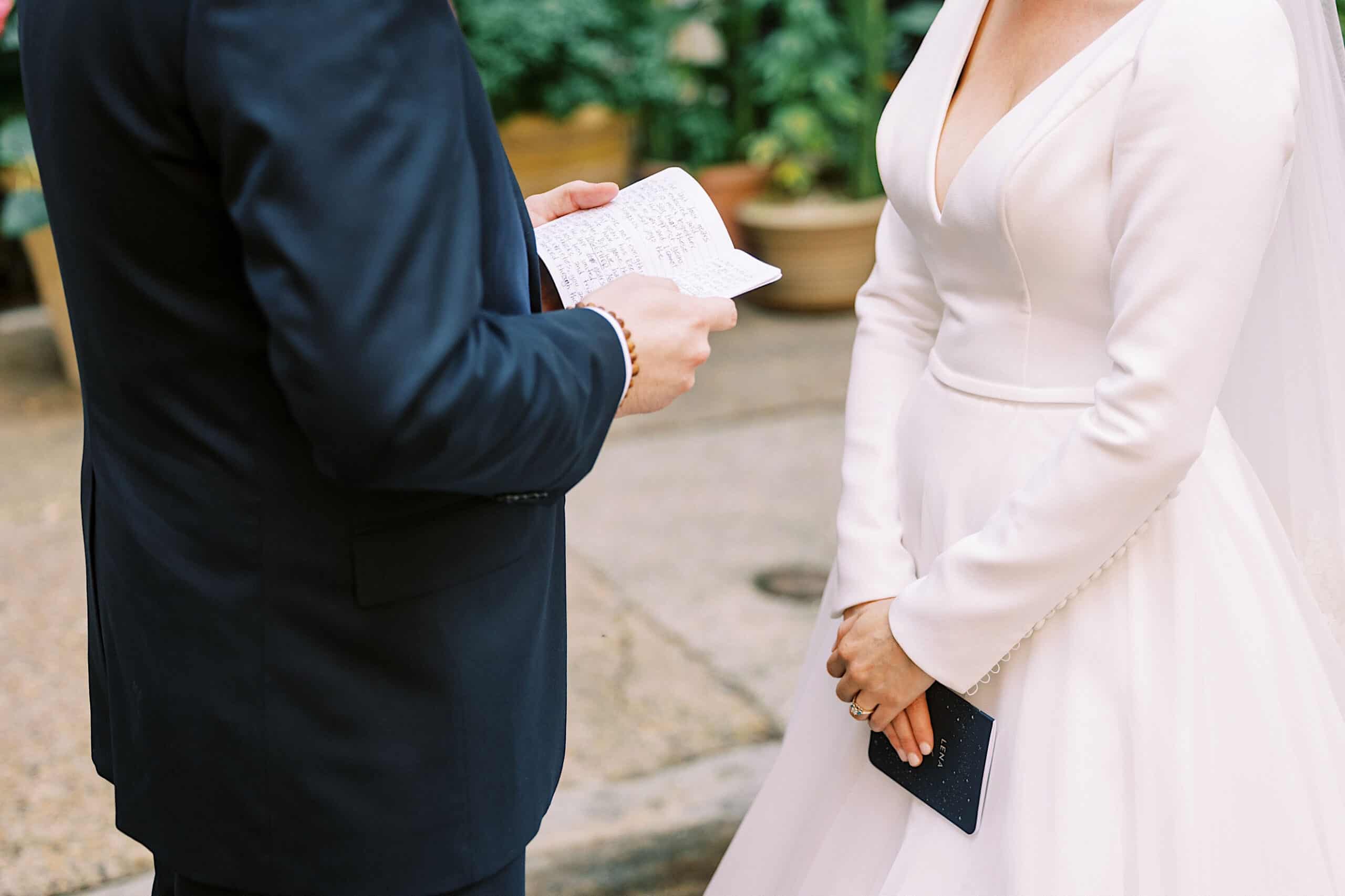 A bride and groom stand facing each other outdoors at their Franklin Institute wedding; the groom holds handwritten vows, while the bride wears a white dress and holds a small notebook.