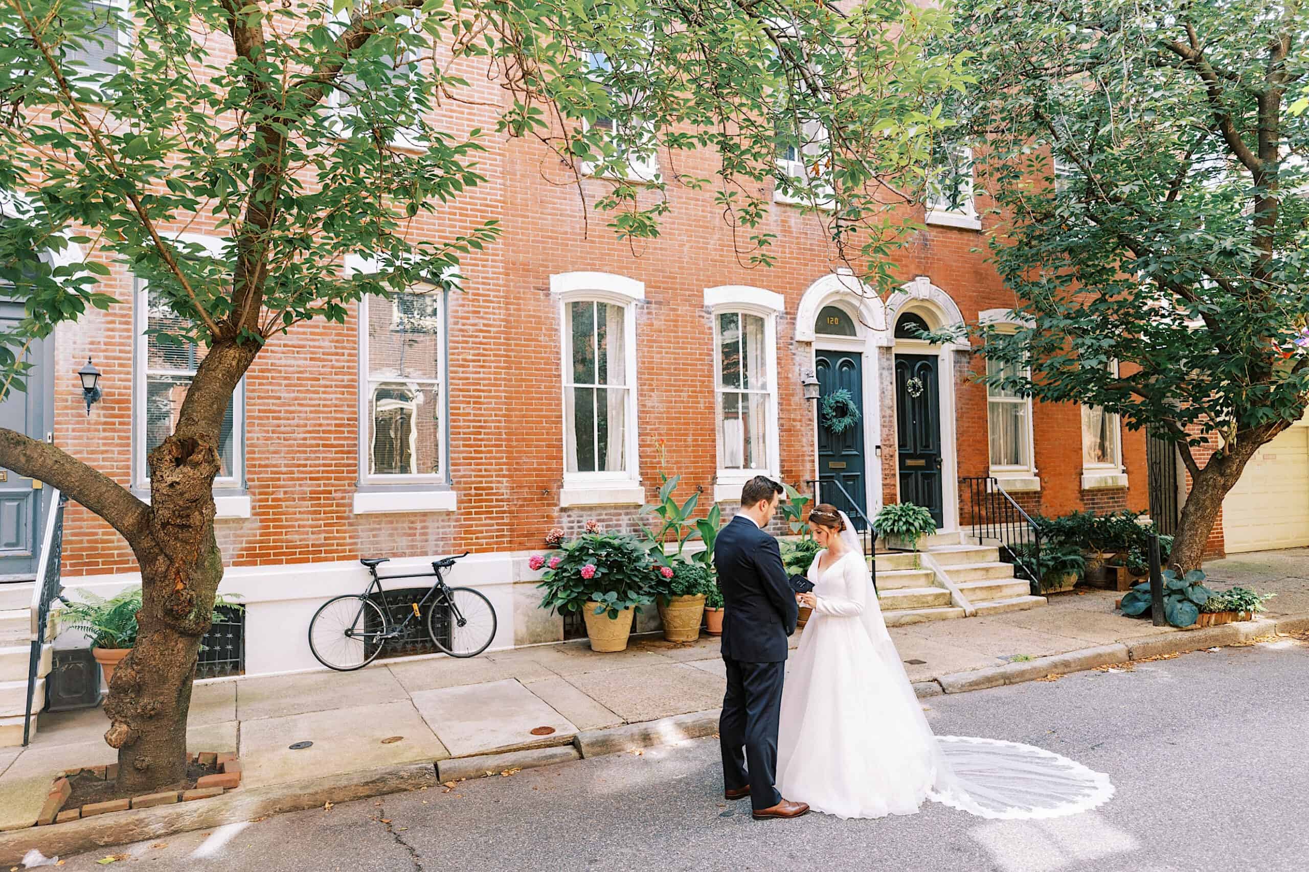 A bride and groom stand facing each other on a quiet tree-lined street in front of a brick townhouse with potted plants and a bicycle, capturing an intimate moment before their Franklin Institute wedding.