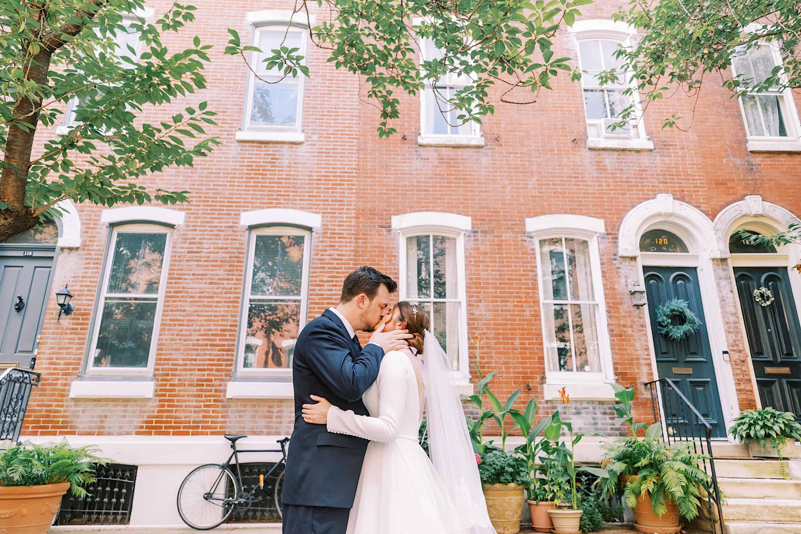 A bride and groom embrace and kiss on a sidewalk in front of a brick townhouse, surrounded by potted plants and a parked bicycle after their Franklin Institute wedding.
