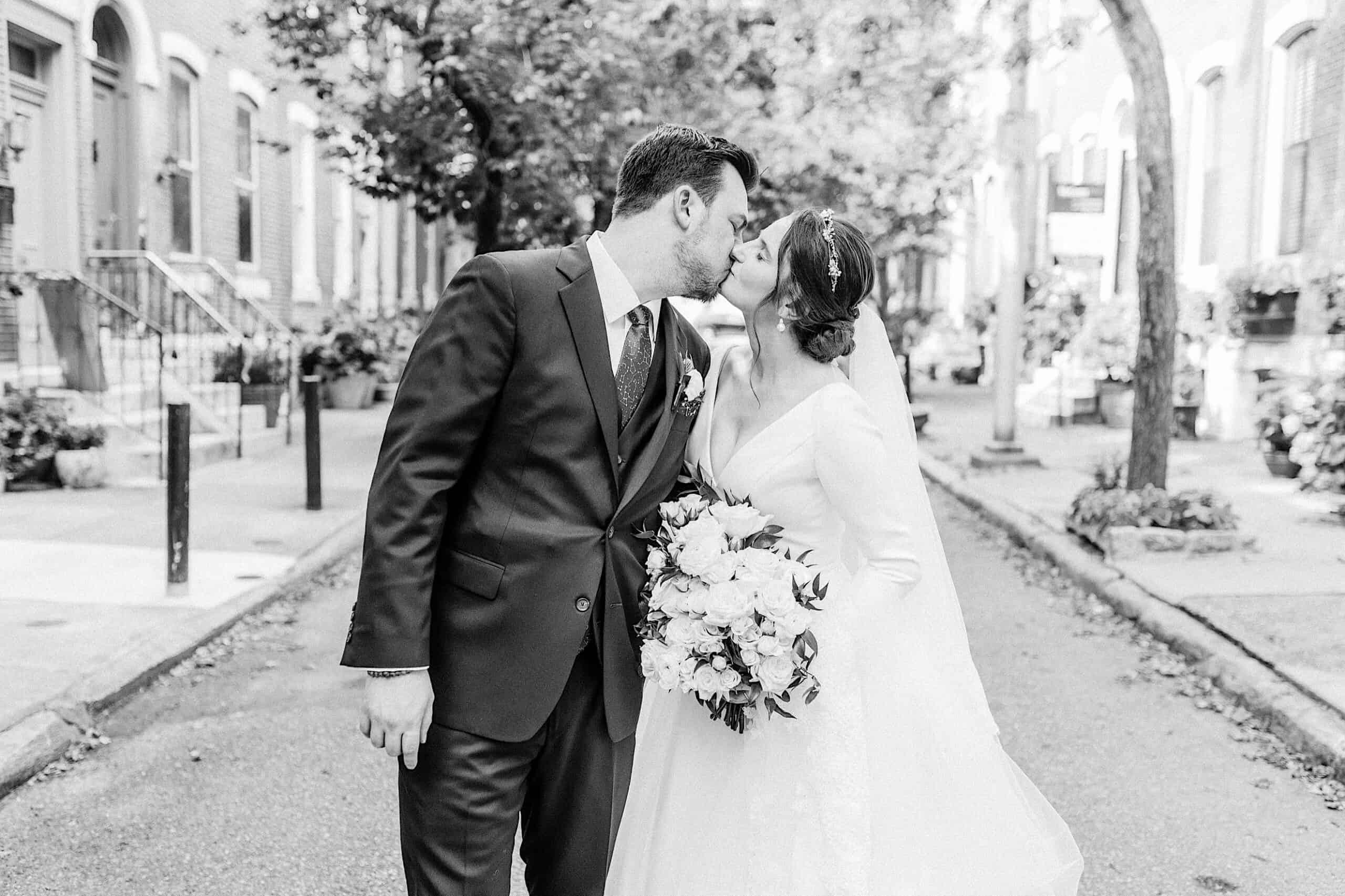 A bride and groom share a kiss on a city street, the bride holding a bouquet of flowers; this timeless black and white moment captures the romance of their Franklin Institute wedding.