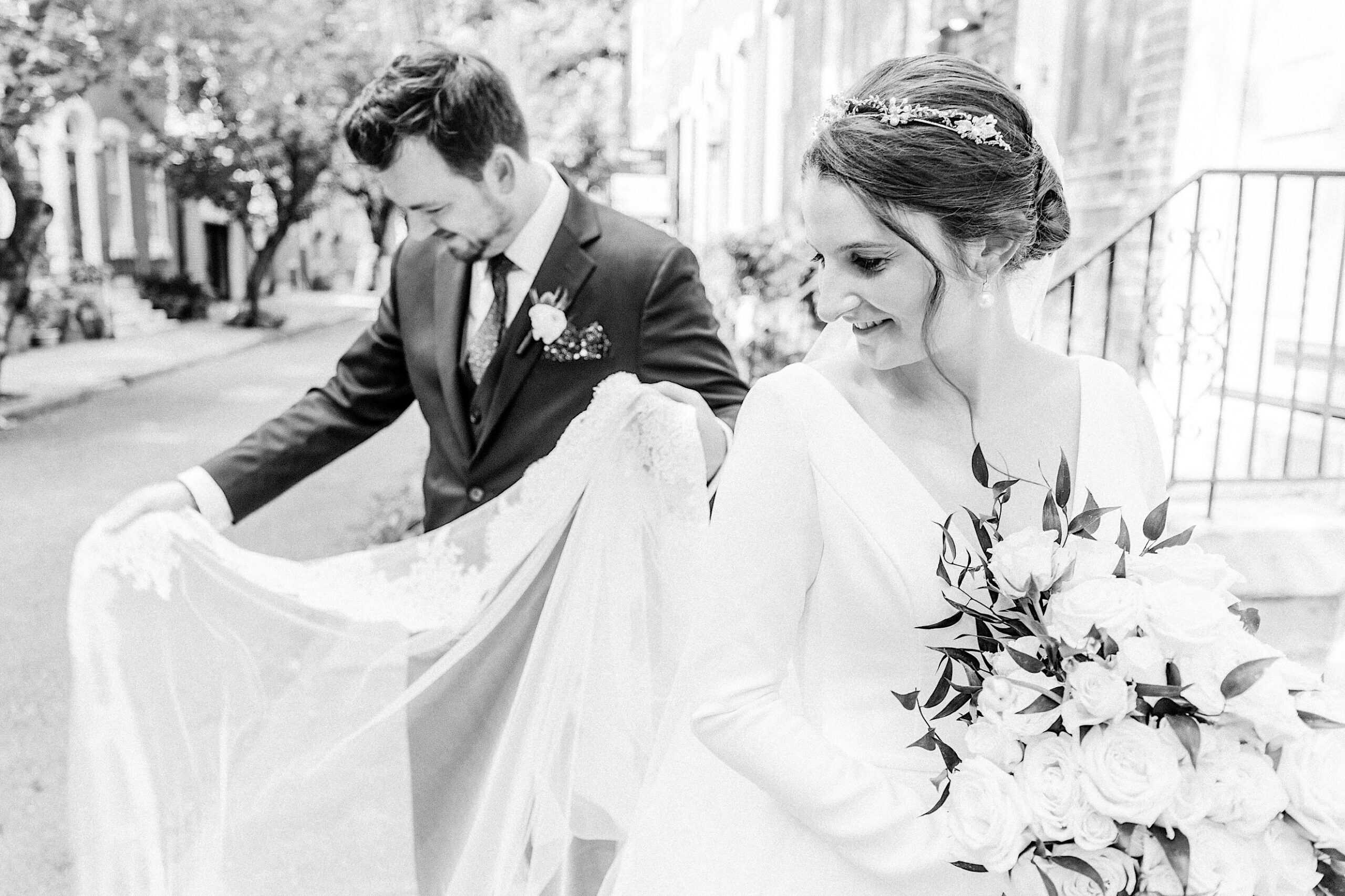 A groom holds the bride's lace veil as she smiles and holds a bouquet of white roses; both are dressed in wedding attire on a city street after their beautiful Franklin Institute wedding.