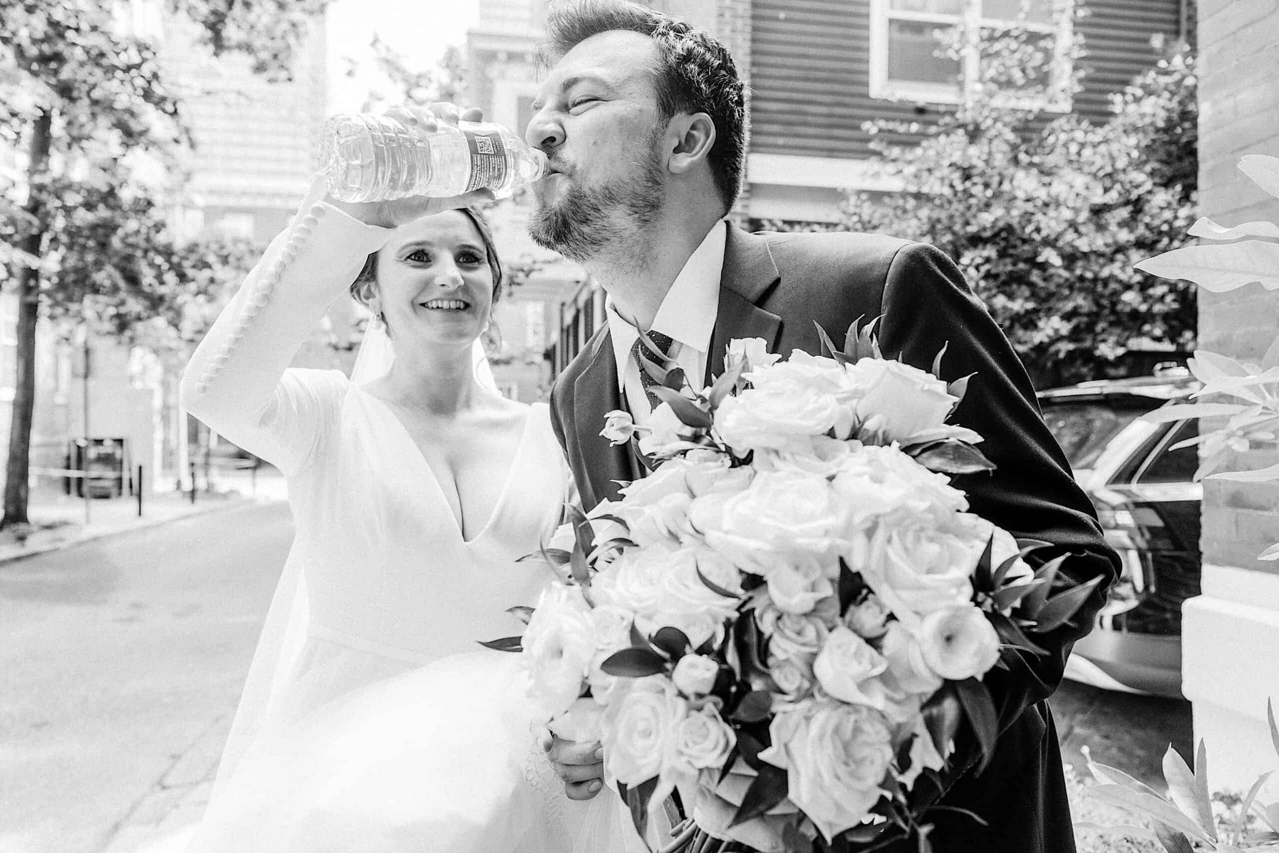 A bride holds a bouquet and helps a groom in a suit drink water from a bottle on a city street, captured in black and white after their Franklin Institute wedding.