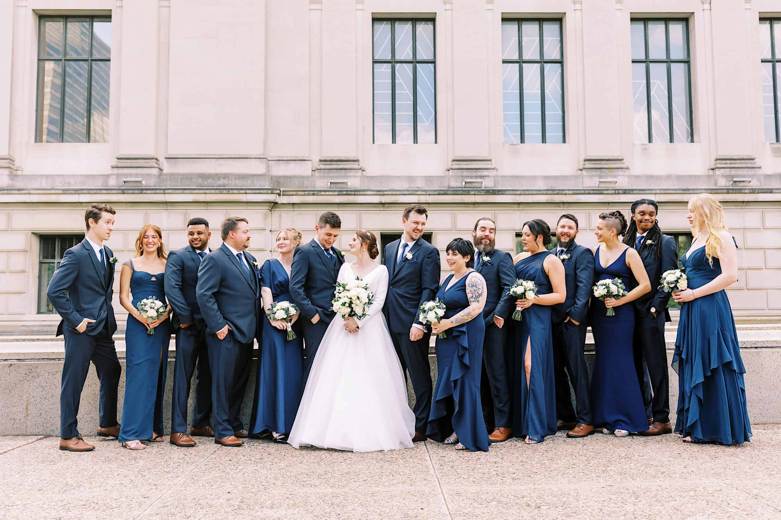 A bride and groom stand with their wedding party, all dressed in navy blue and white attire, posing outdoors in front of the iconic stone facade of the Franklin Institute wedding venue.