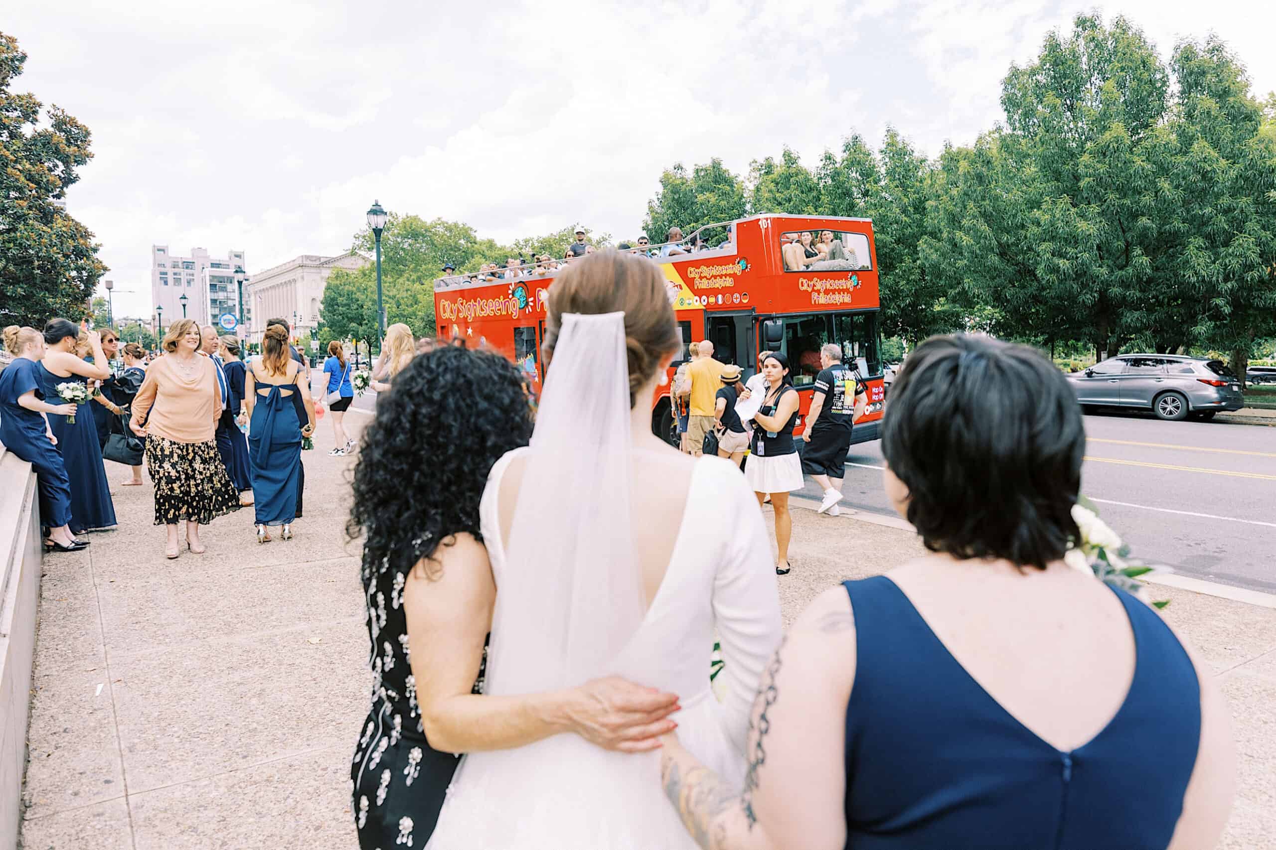 A bride in a white dress and veil stands with two women, facing a red double-decker tour bus on a city street near the Franklin Institute wedding venue, with people gathered around.