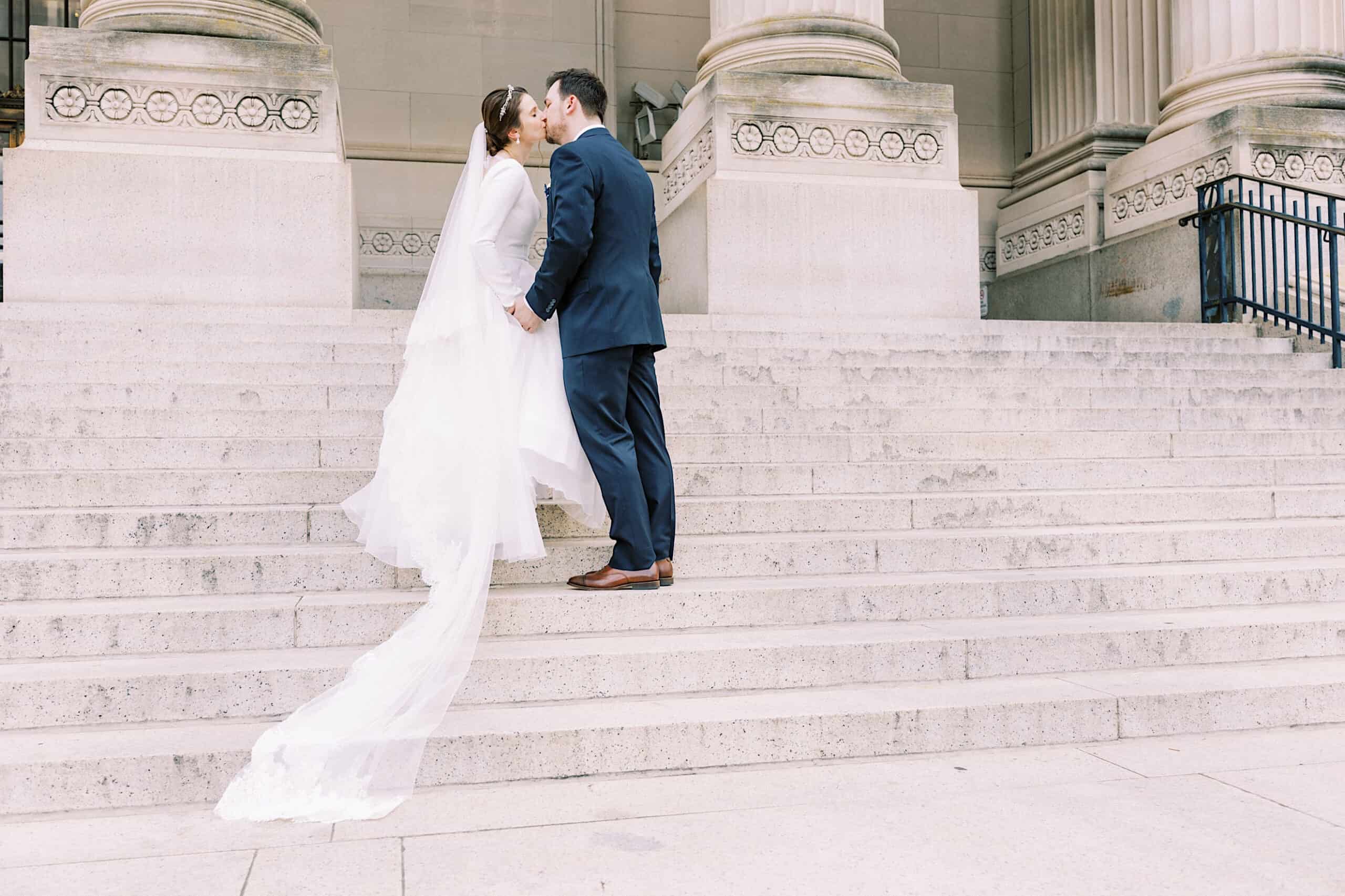A bride and groom stand on stone steps, facing each other and holding hands, in front of the grand columns of the Franklin Institute wedding venue.