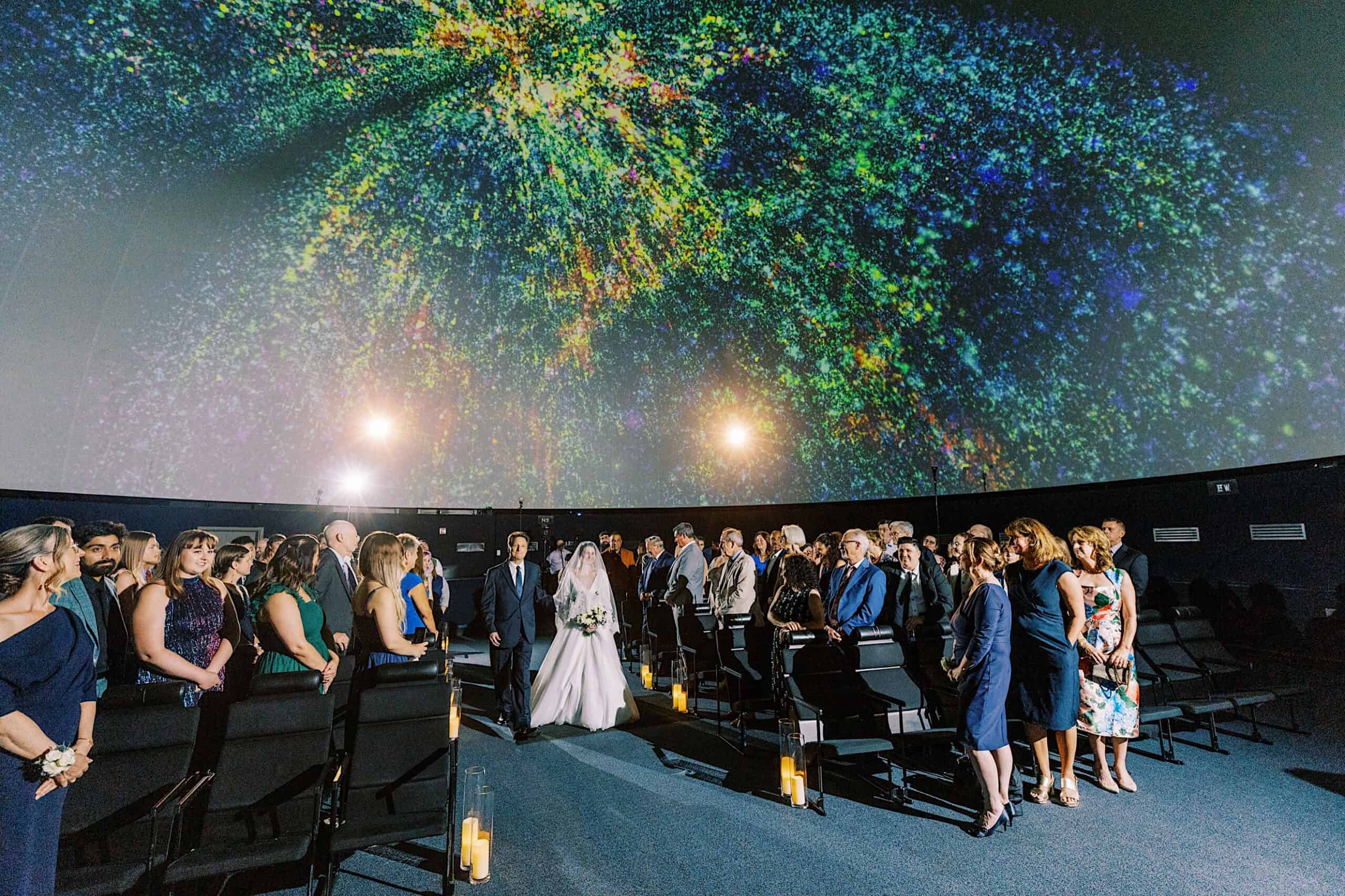 A bride and groom walk down the aisle in a dark, dome-shaped Franklin Institute wedding venue with colorful lights projected on the ceiling as guests stand on either side.