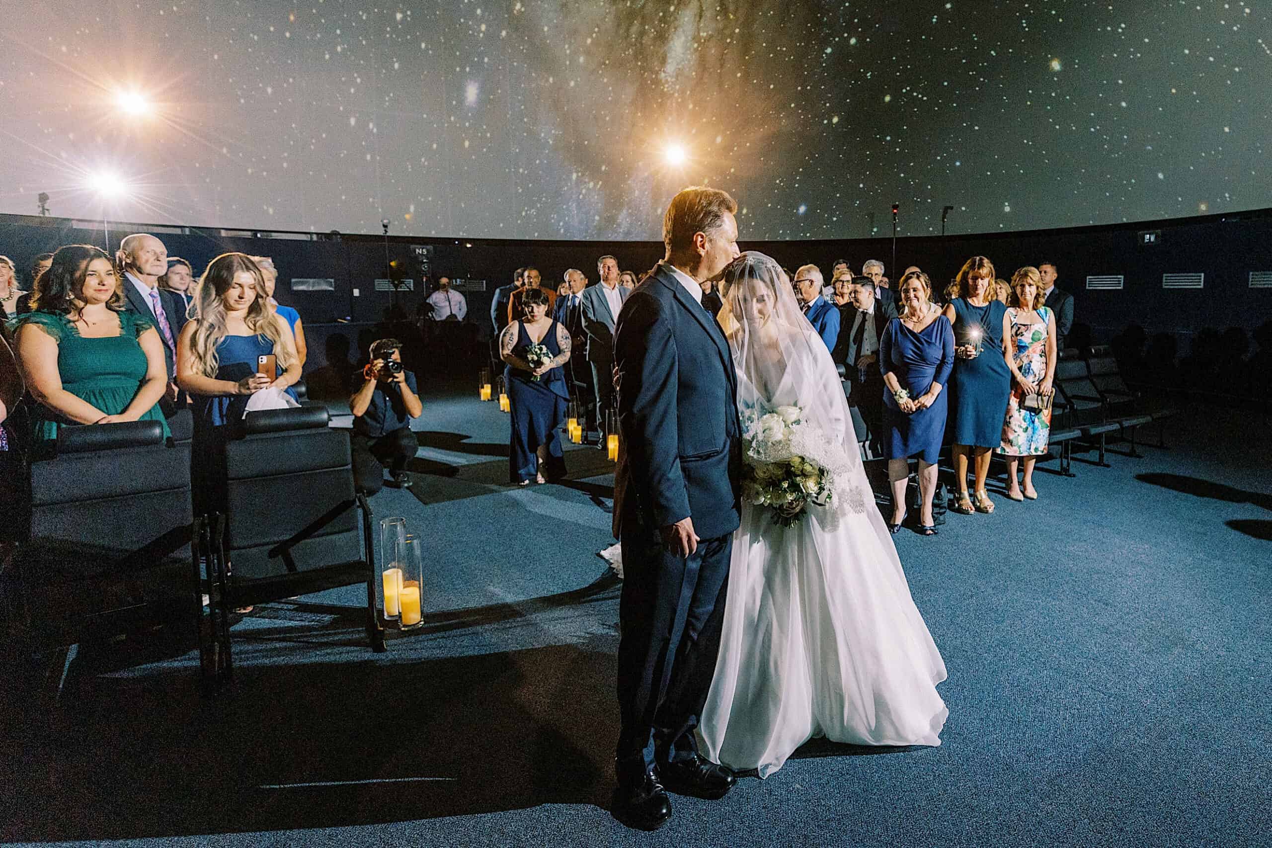 A bride in a white gown and veil stands with a man, likely her father, before seated guests under a projected starry night sky at a Franklin Institute wedding ceremony.