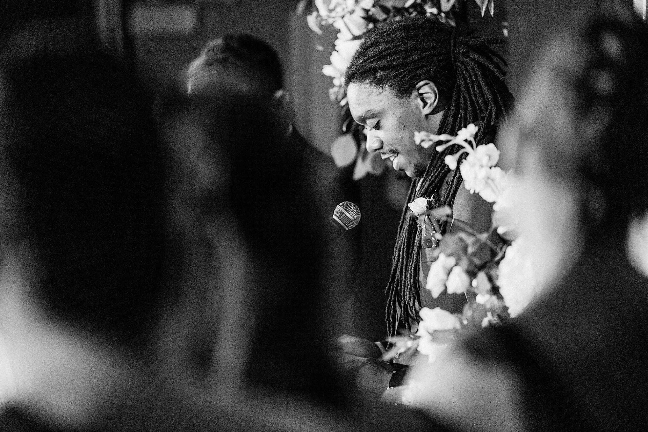 A man with long dreadlocks stands at a microphone, surrounded by flowers, speaking or reading at what appears to be a Franklin Institute wedding. The image is in black and white.