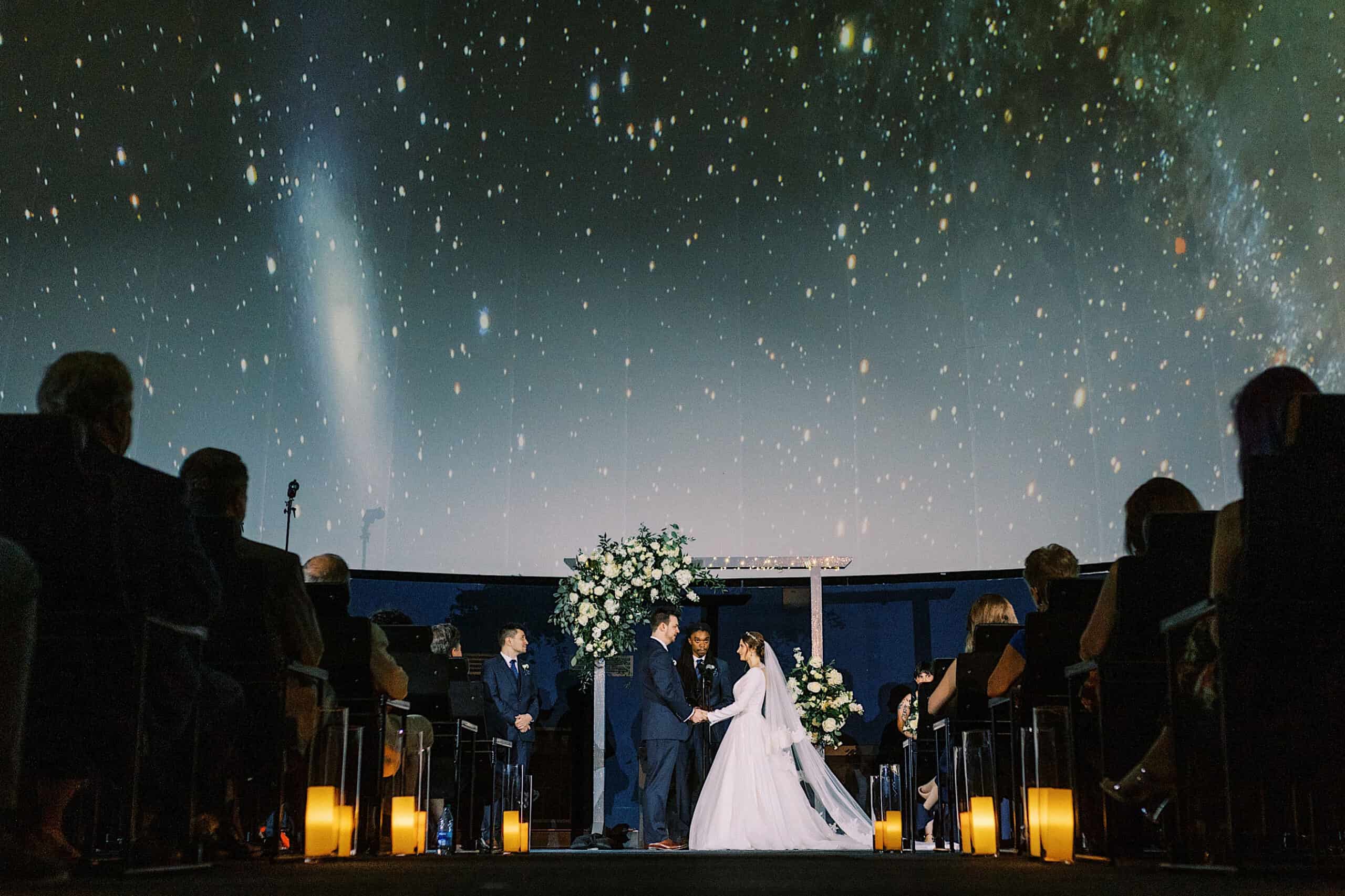 A bride and groom stand at the altar in a darkened room with a starry sky projected above them at their Franklin Institute wedding, surrounded by floral arrangements and seated guests.