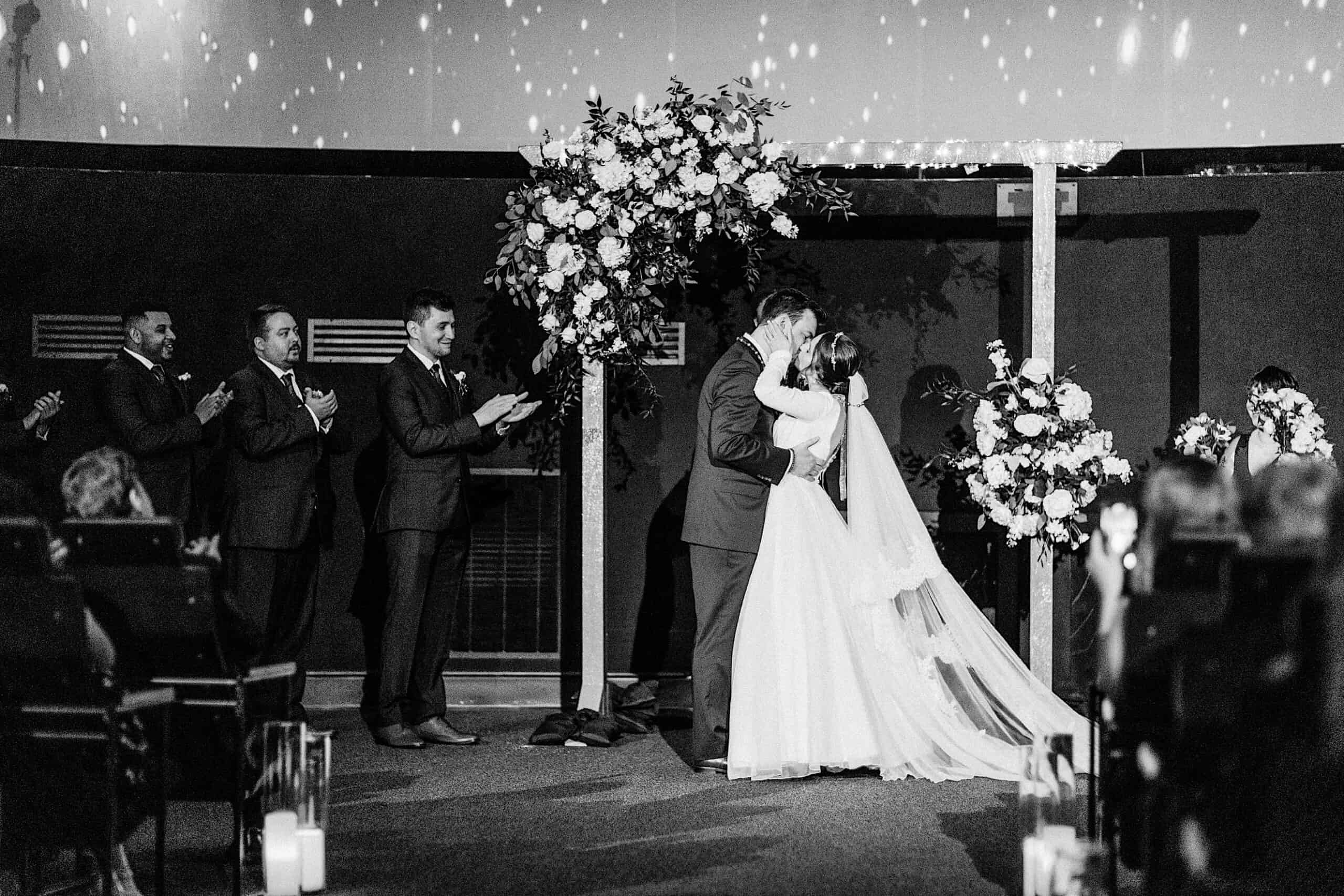 A bride and groom share a kiss under a floral arch during their Franklin Institute wedding ceremony as guests and groomsmen look on and applaud.