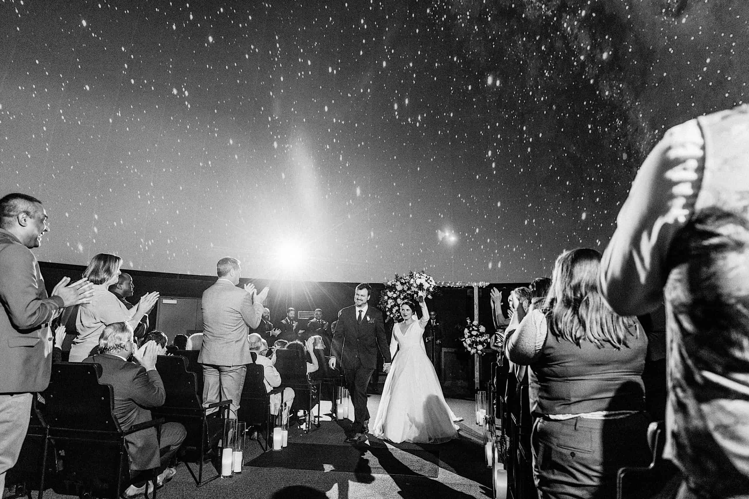 A bride and groom walk down the aisle as guests applaud, with a starry night sky overhead in this black and white outdoor Franklin Institute wedding scene.