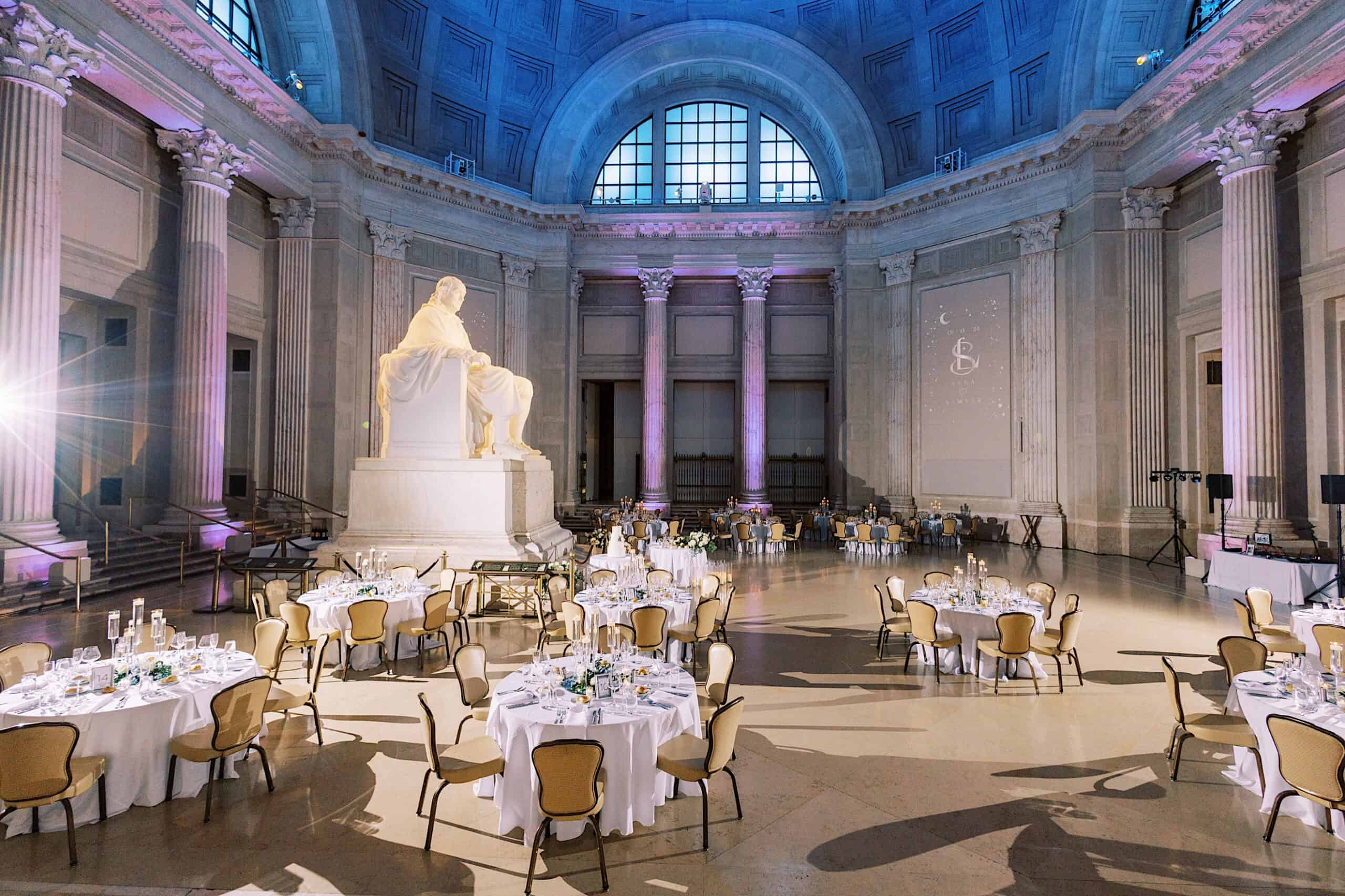 Large, elegant hall with round tables set for a Franklin Institute wedding, featuring a prominent white statue and tall columns under a high arched ceiling with blue lighting.
