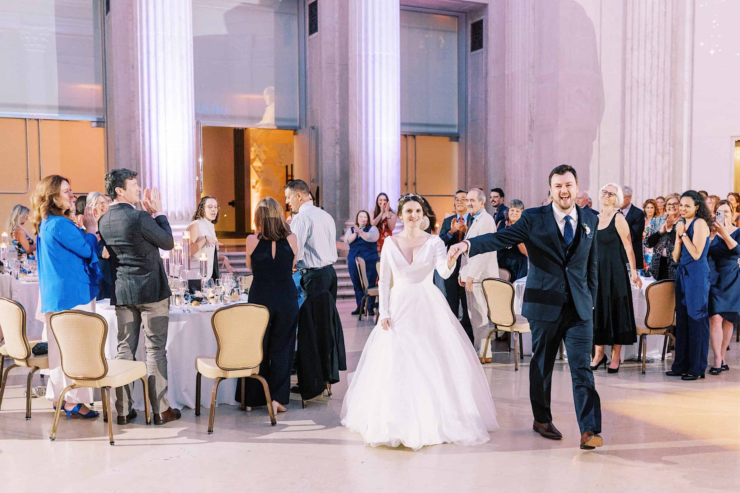 A bride and groom enter the elegant Franklin Institute wedding reception hall as guests stand and applaud.