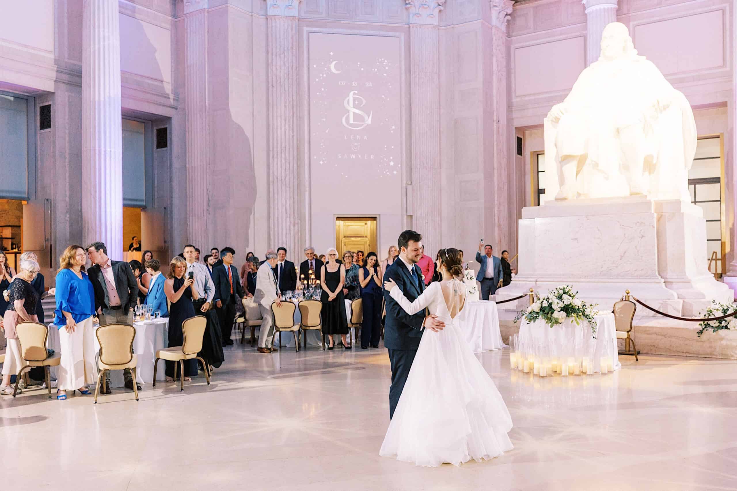 A bride and groom share their first dance in a grand hall at their Franklin Institute wedding, with guests watching and a large statue and wedding monogram projected on the wall in the background.