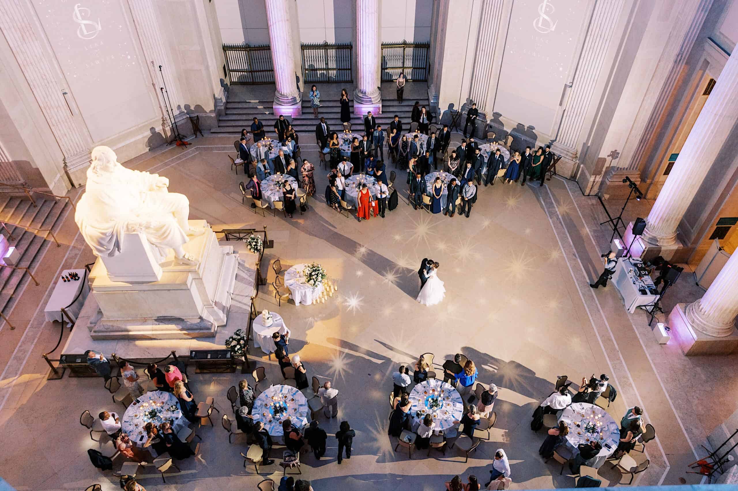 A bride and groom share their first dance in a grand hall at their Franklin Institute wedding, with round tables of seated guests and a large white statue in the foreground.