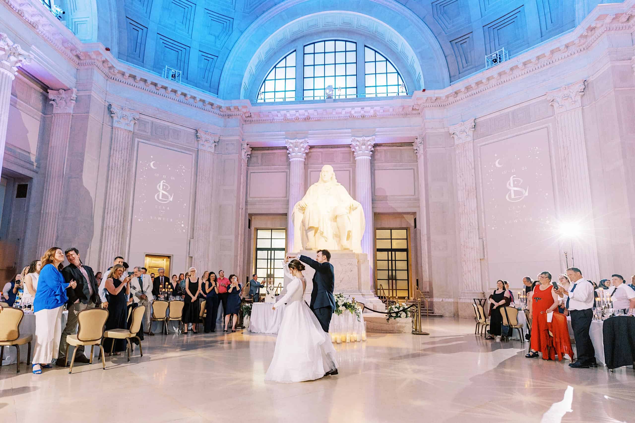 A bride and groom share their first dance in the grand hall of a Franklin Institute wedding, with a large seated statue and wedding guests watching.