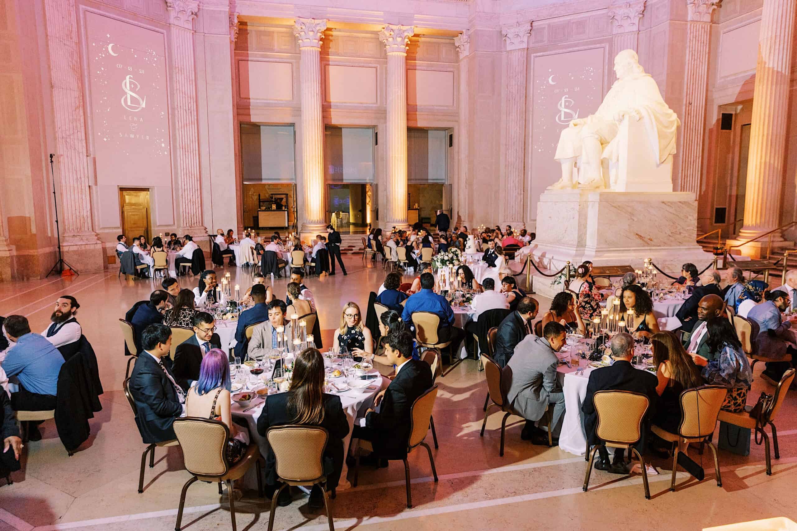 Guests seated at round tables enjoy dinner in a grand hall with tall columns and a large white statue, under warm lighting—a stunning setting for a Franklin Institute wedding.