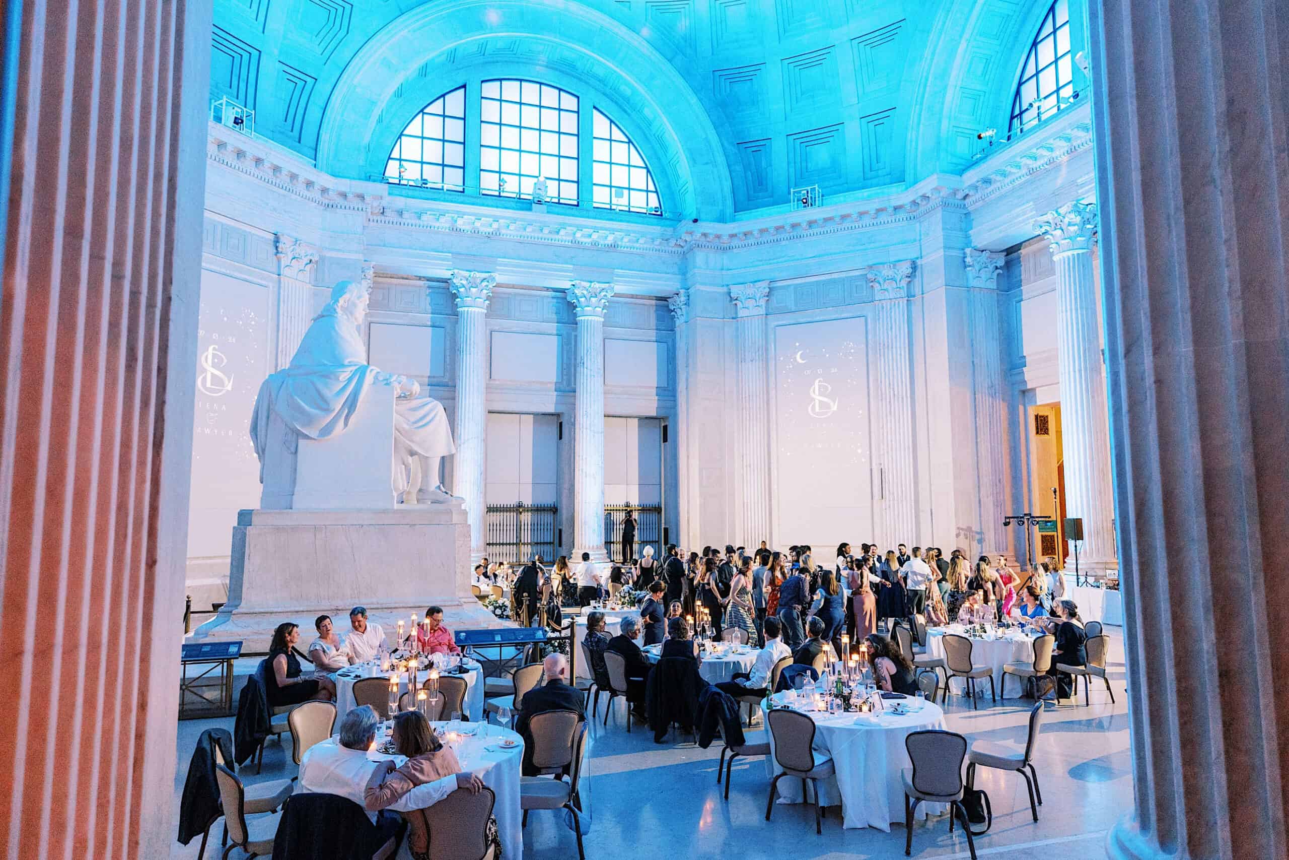 Guests seated at round tables in a grand hall with high arched ceilings, tall columns, blue lighting, and a large statue at the front enjoy the elegance of a Franklin Institute wedding.