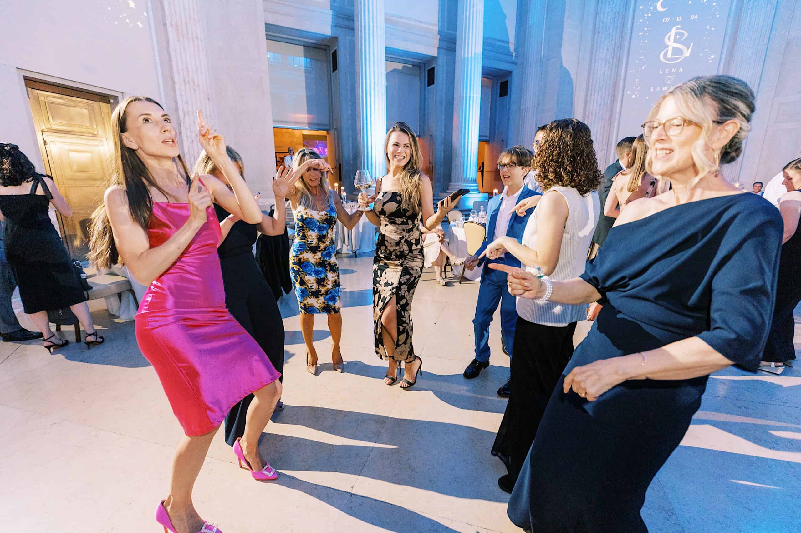 A group of women in formal attire dance and smile together at a Franklin Institute wedding, surrounded by high ceilings and white columns at the elegant indoor event.