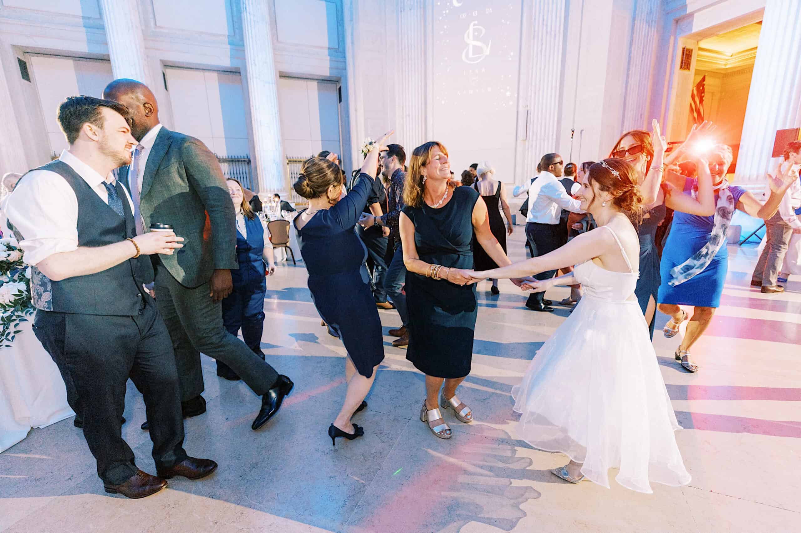 People in formal attire dance and celebrate in a large, elegant hall with tall columns and high ceilings during a Franklin Institute wedding reception.