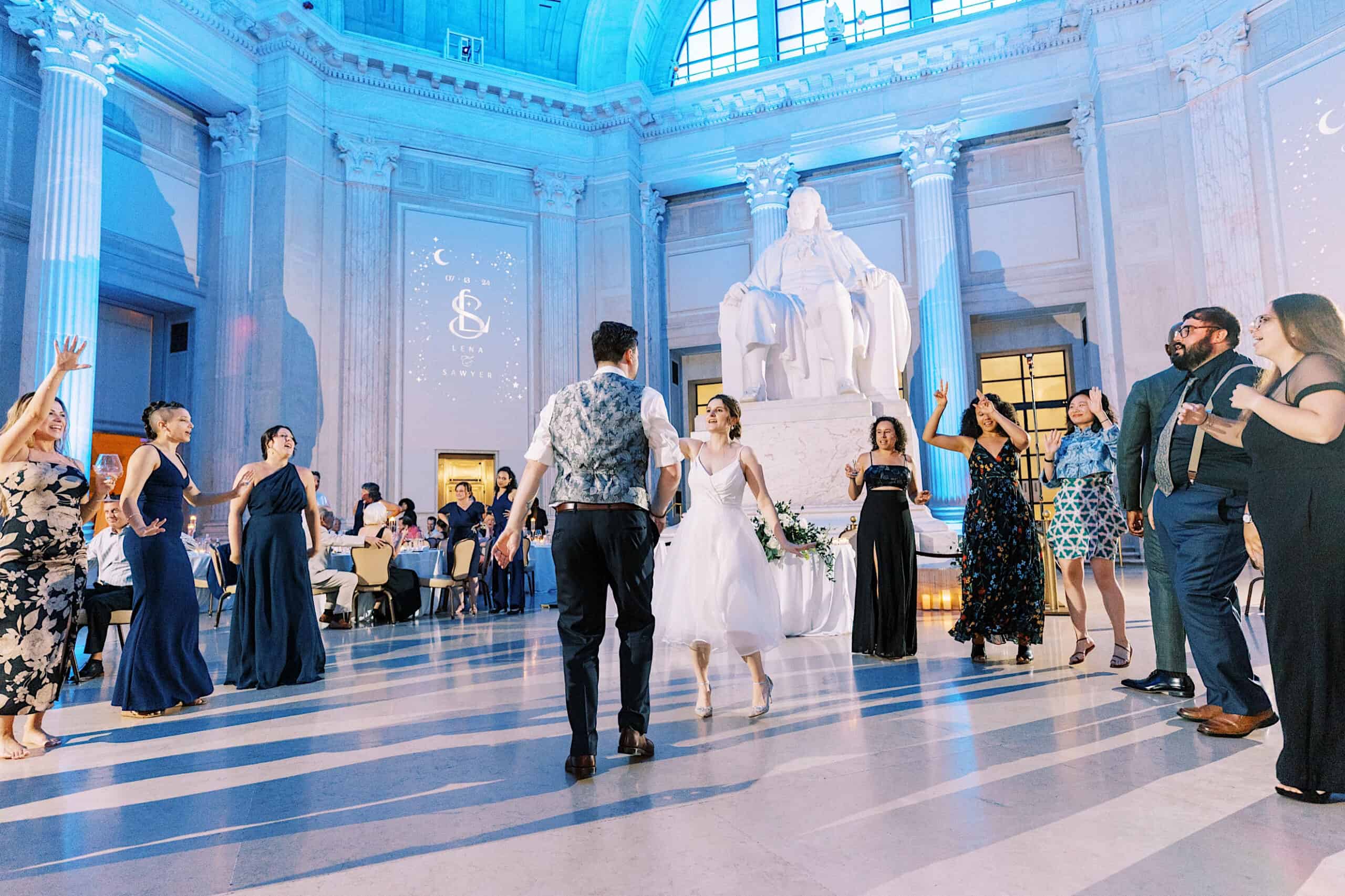 A bride and groom dance in a grand hall with tall columns and a large white statue at their elegant Franklin Institute wedding, surrounded by guests in formal attire.