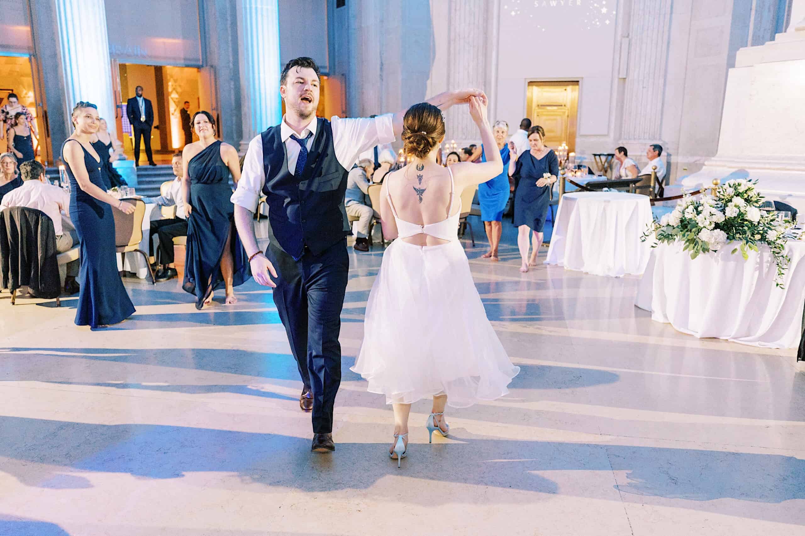 A man in a dark vest dances with a woman in a white dress at their Franklin Institute wedding, surrounded by guests seated and standing in an elegant, well-lit hall.